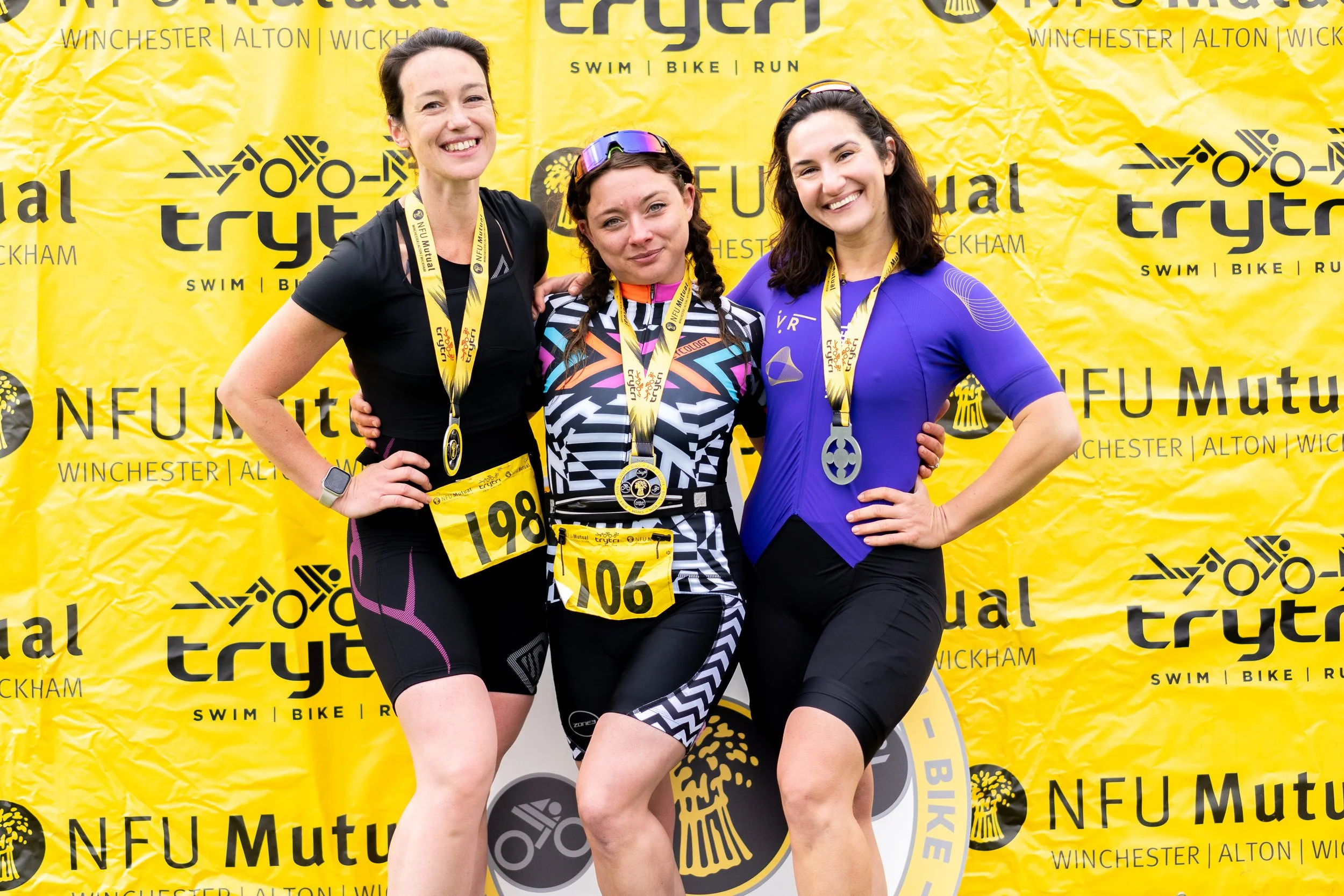Three women standing together, wearing athletic gear and medals, smiling at a race event with a yellow backdrop that reads 'NFU Mutual Winchester Alton Wickham Triathlon' and lists swim, bike, run categories.