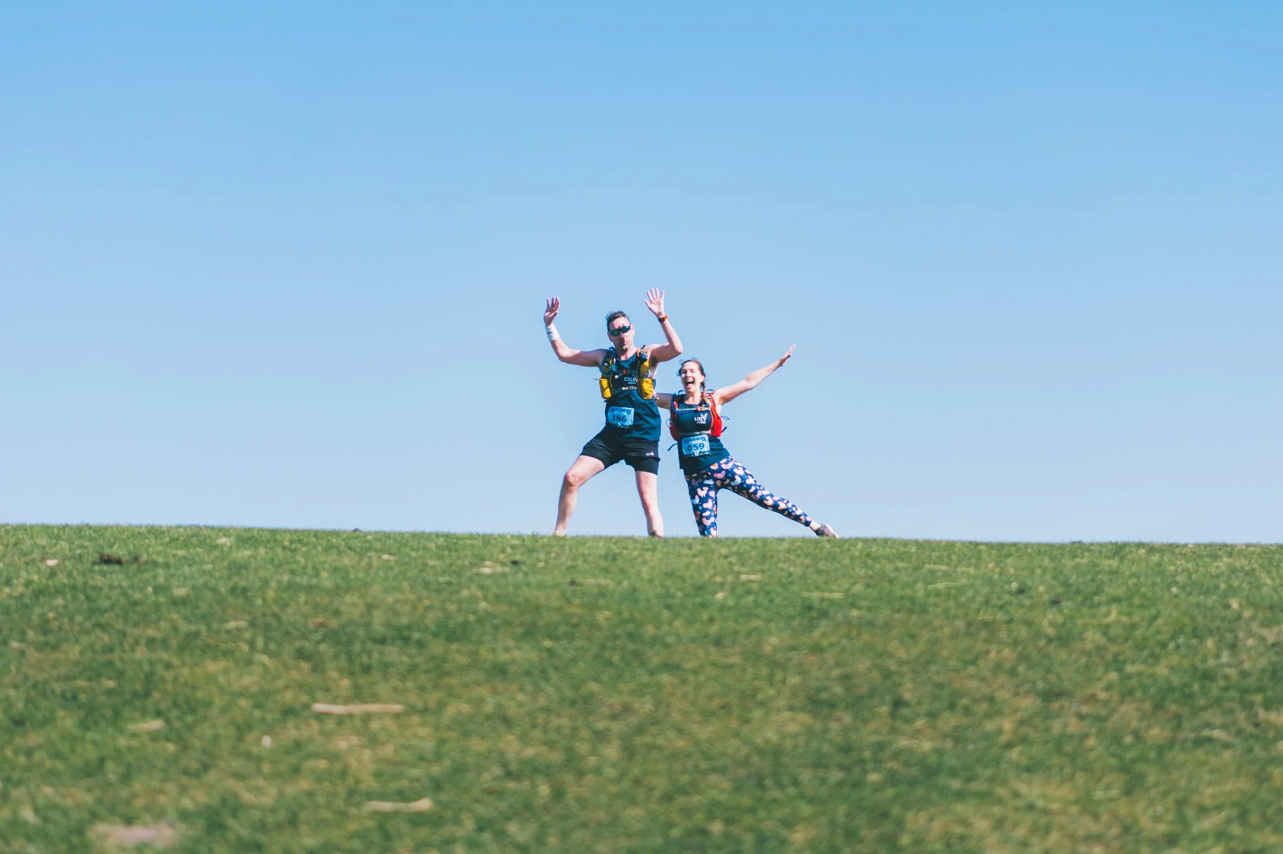 Two runners celebrating on a grassy hill against a clear blue sky.