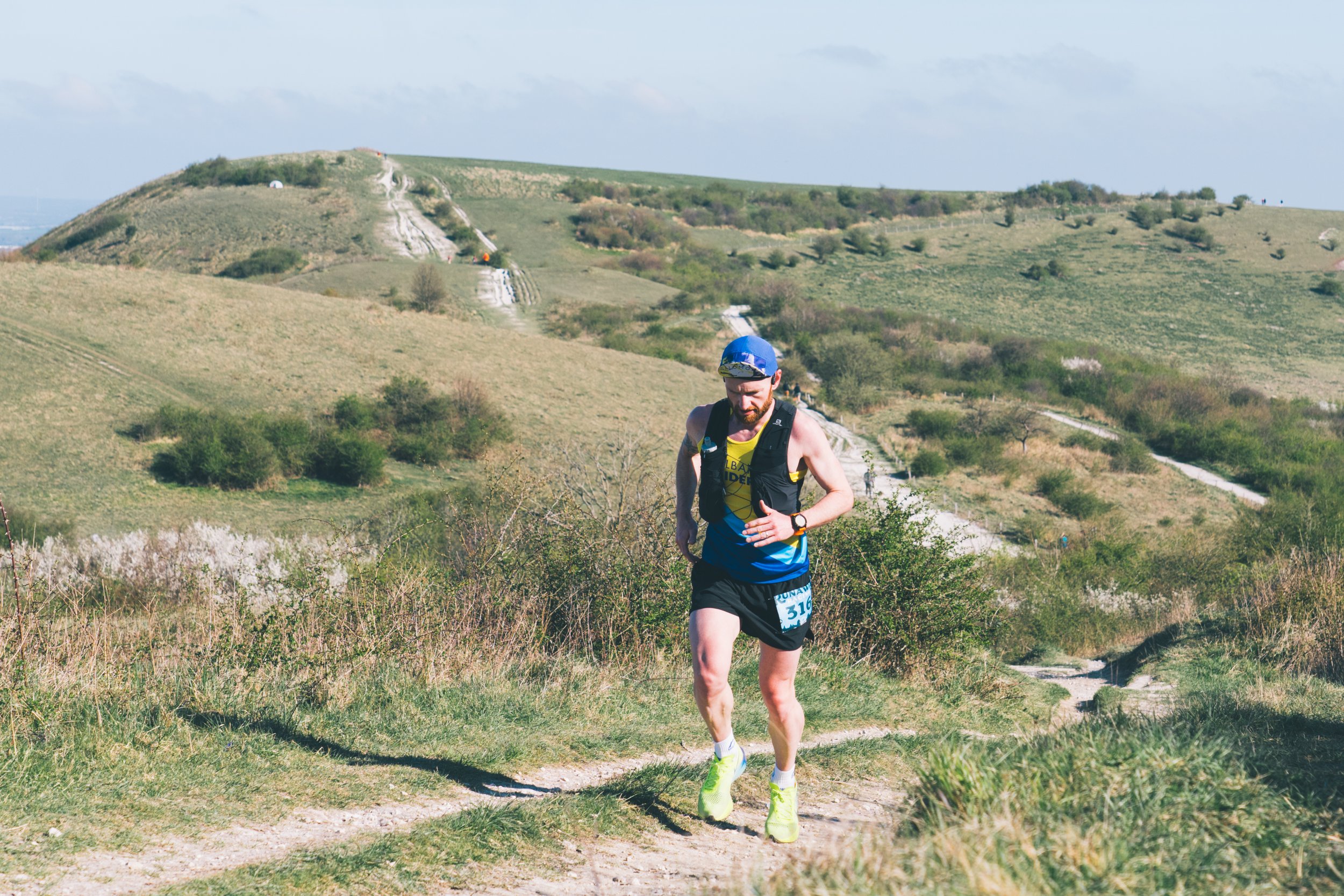 A male runner in a blue shirt, black shorts, wearing a blue helmet and a black running vest, running on a trail through a hilly, grassy landscape.
