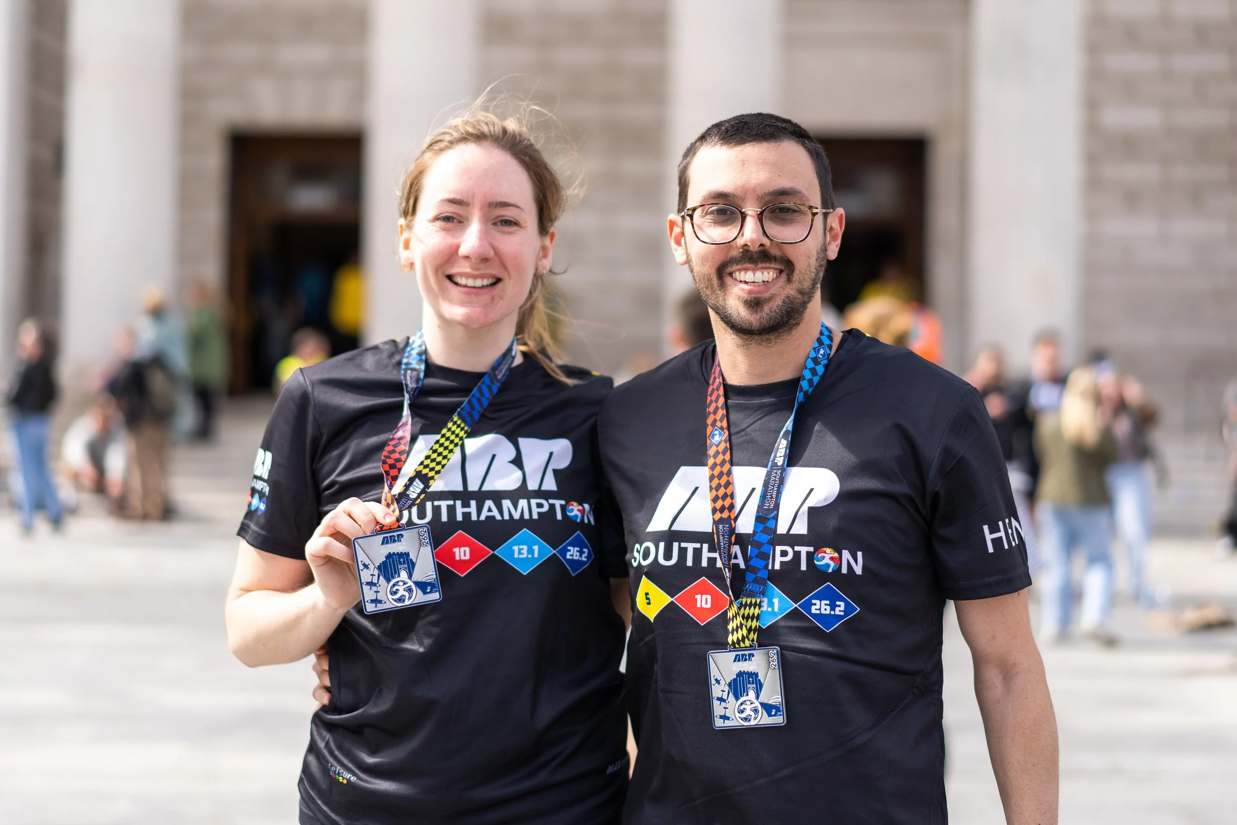 Two smiling marathon runners, a woman and a man, wearing black race shirts with 'ABP SOUTHAMPTON' logo and medals around their necks, standing in front of a building with steps.