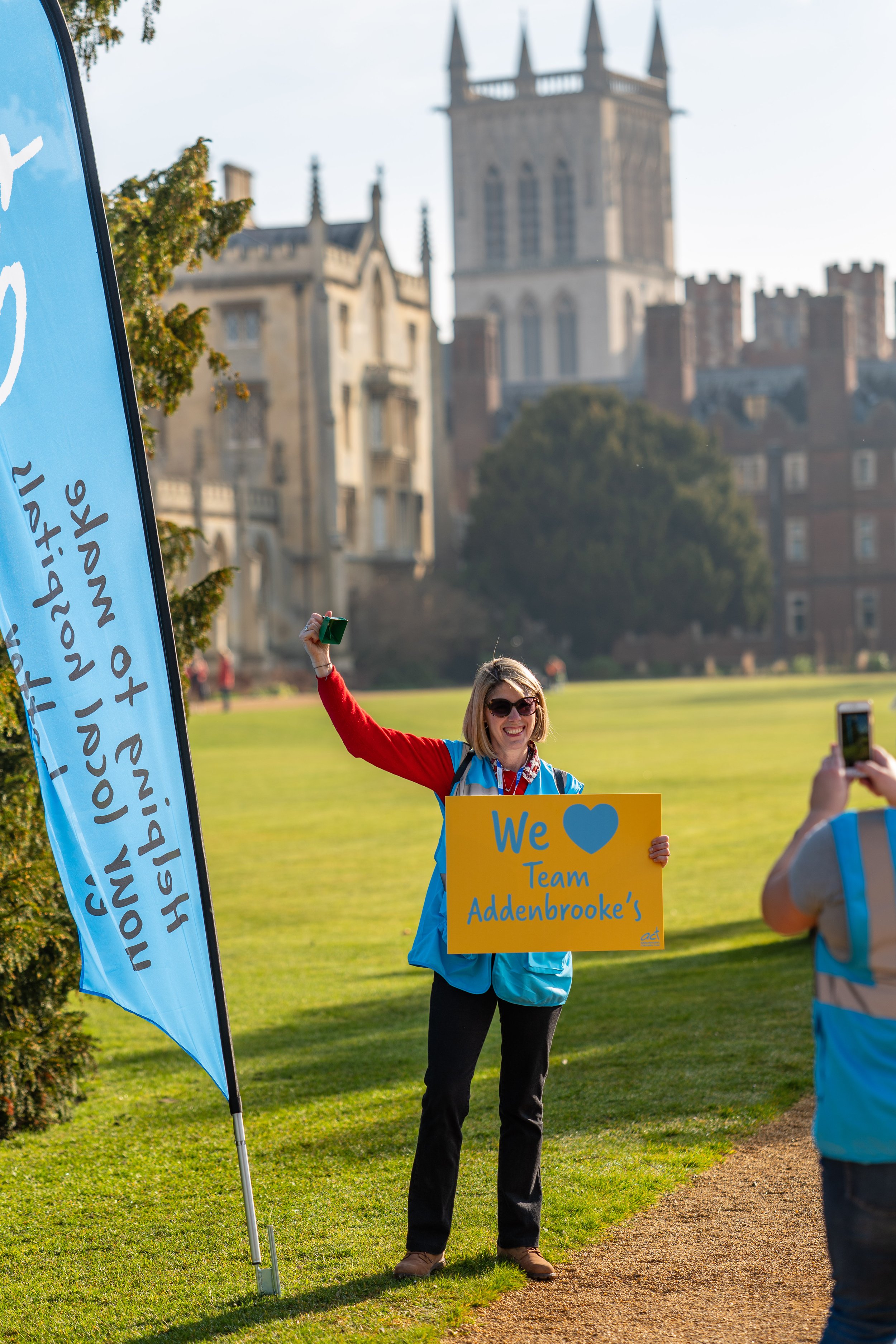 A woman in sunglasses smiling and holding a yellow sign that reads 'We ♥ Team Addenbrooke's,' standing on a grassy area with historic buildings in the background. Another person is taking her picture.