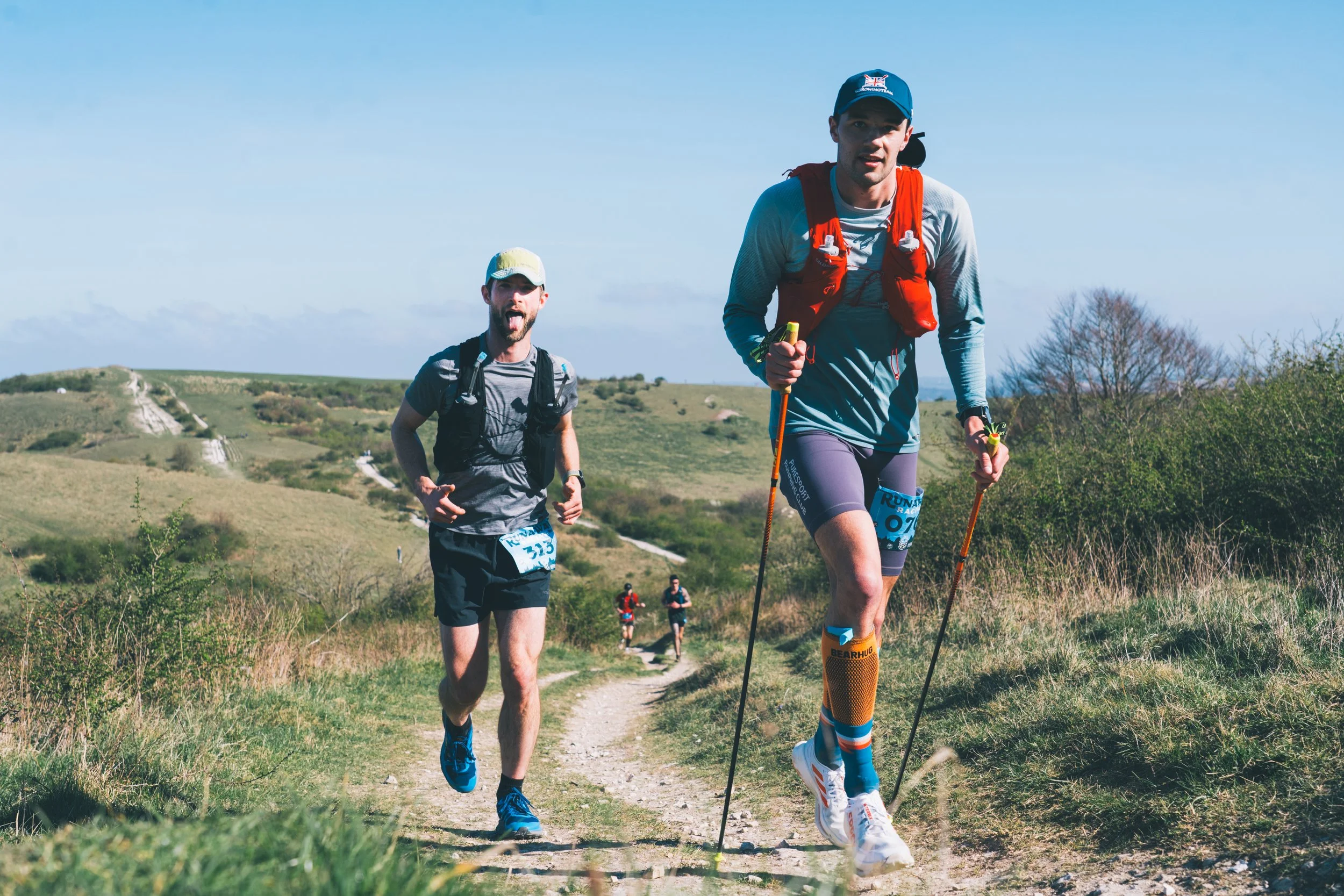 Two male trail runners outdoors on a dirt trail in a hilly landscape, with three other runners in the background, under a partly cloudy sky.