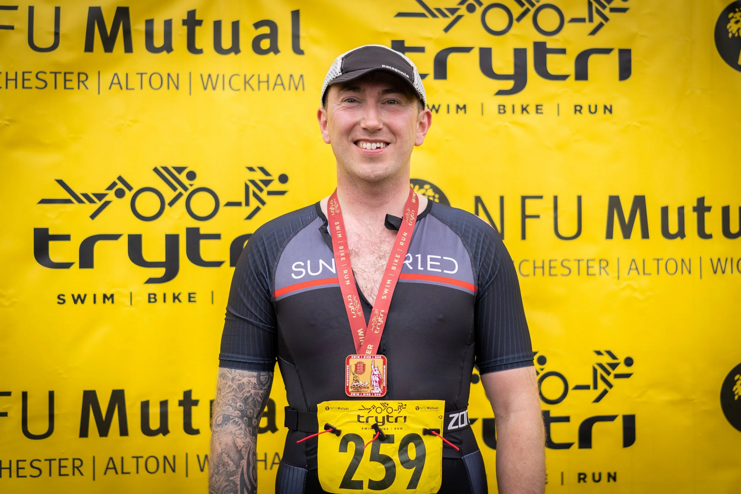 A smiling man wearing a black and gray athletic shirt with red accents, a race bib numbered 259, and a medal around his neck, standing in front of a yellow backdrop with black text and logos related to a triathlon event.