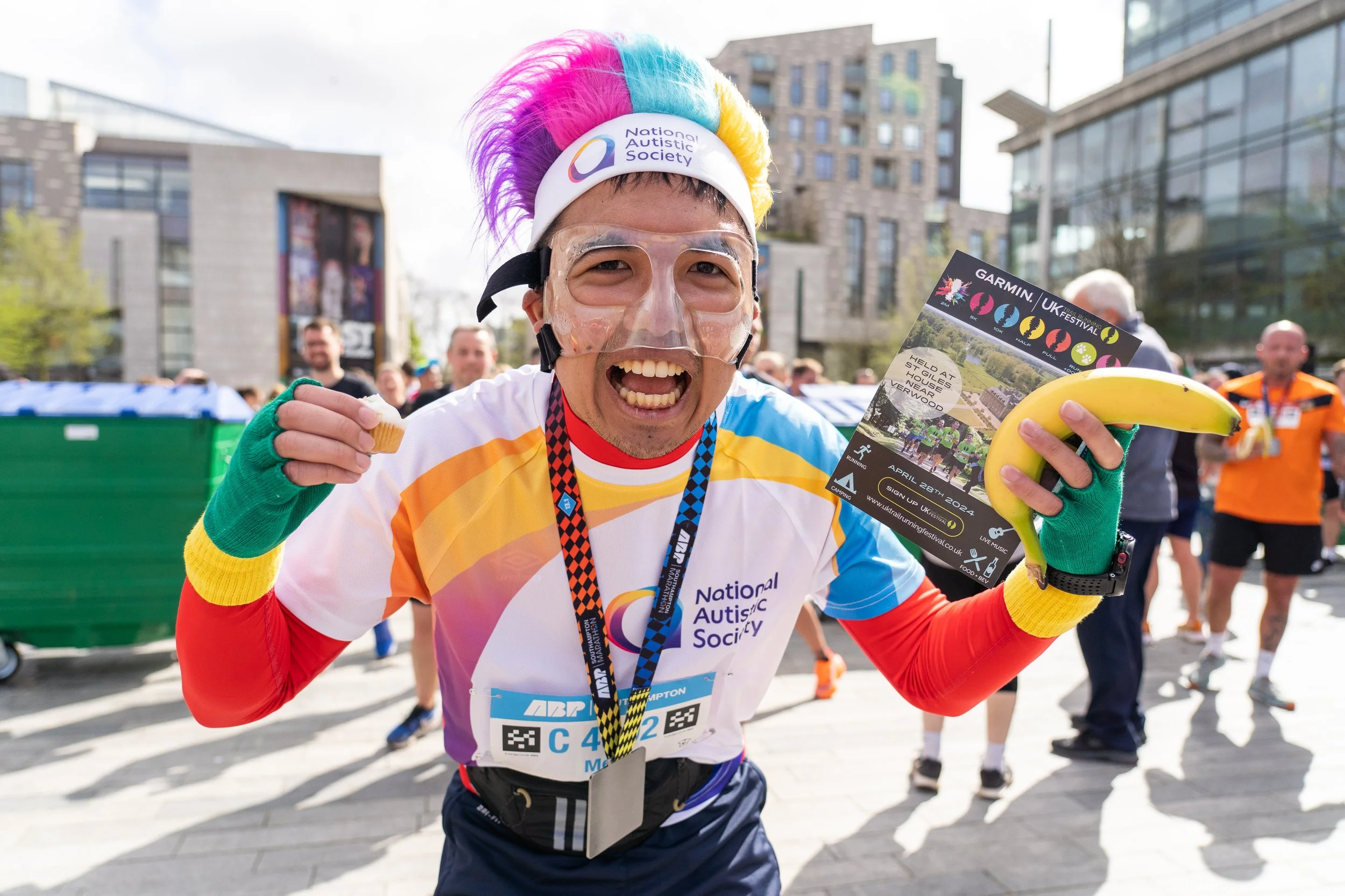 A man celebrating after a race, wearing a colorful rainbow wig, a face shield, a National Autistic Society t-shirt, and green gloves, holding a banana, a cupcake, and a race flyer, with other runners and buildings in the background.