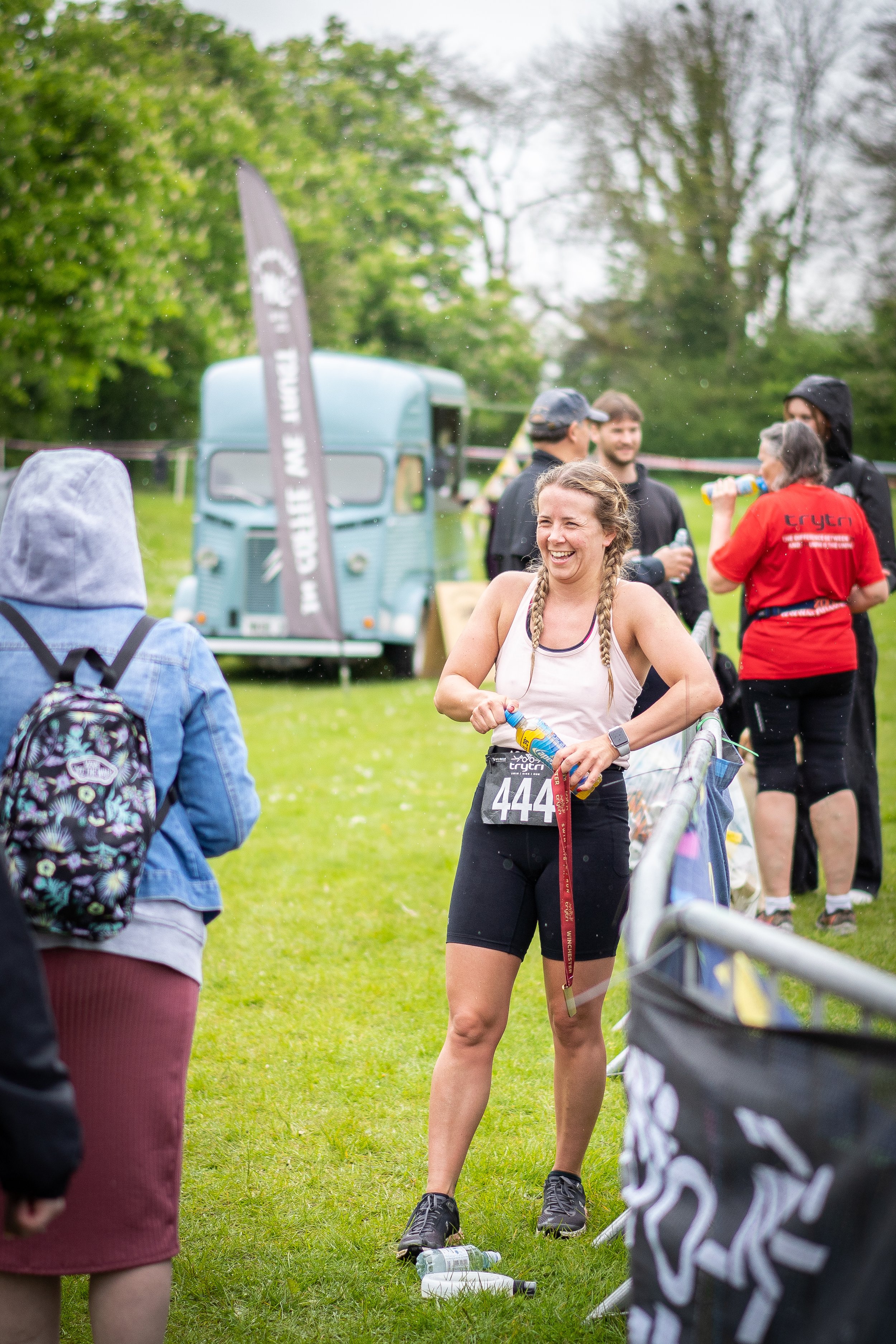 A woman with bib number 444 is smiling and holding a water bottle after a race, with other runners and a vintage light blue van in the background.
