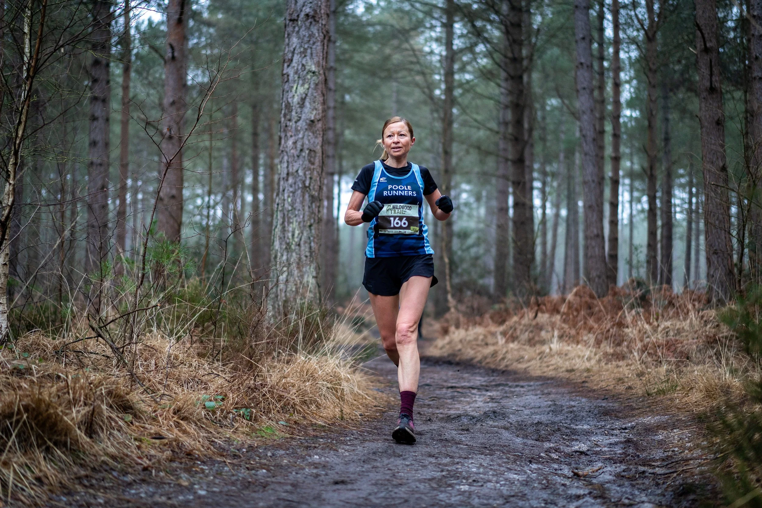 A woman running on a dirt trail through a forest of tall pine trees during a race, wearing a blue and black athletic outfit with a race bib.