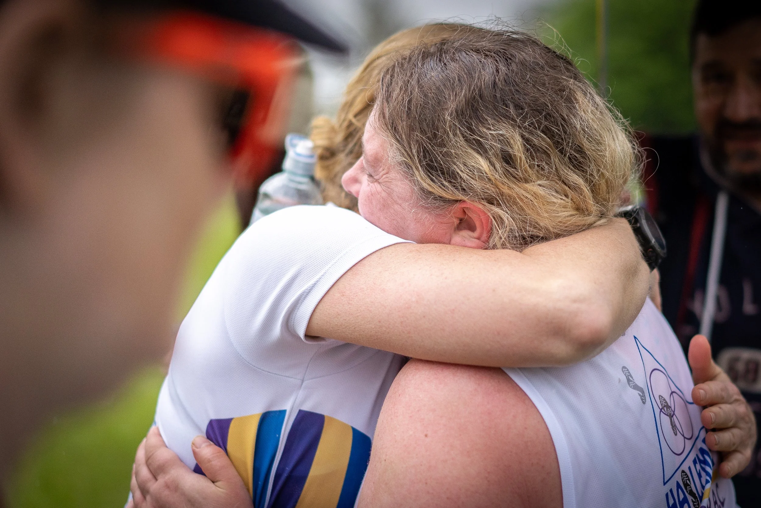 Two women hugging tightly, emotional moment, one wearing a white athletic shirt with a logo on it, outdoor setting with other people in the background.