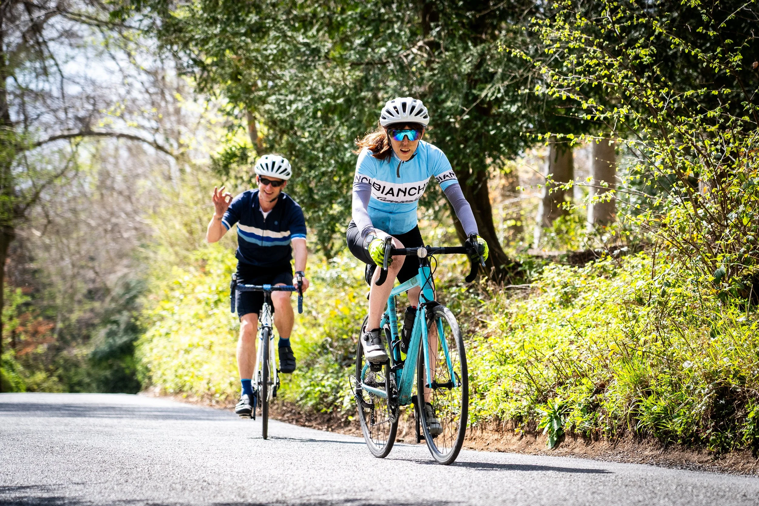 Two cyclists, a woman in front and a man behind, riding on a paved trail through a wooded area with green foliage and sunlight.