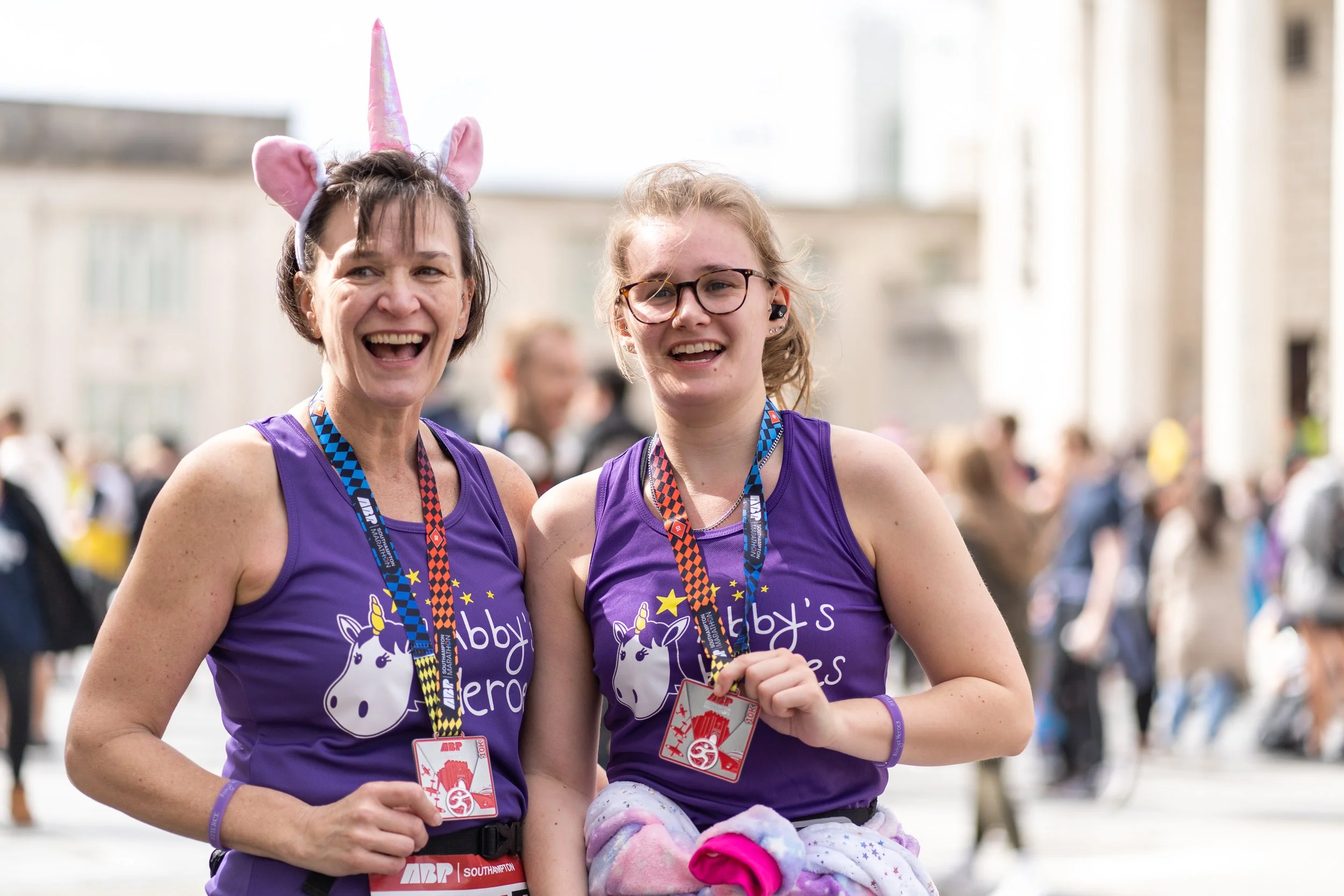 Two women wearing purple unicorn-themed shirts and medals around their necks, smiling after a race, with one wearing unicorn ears and a unicorn horn headband.