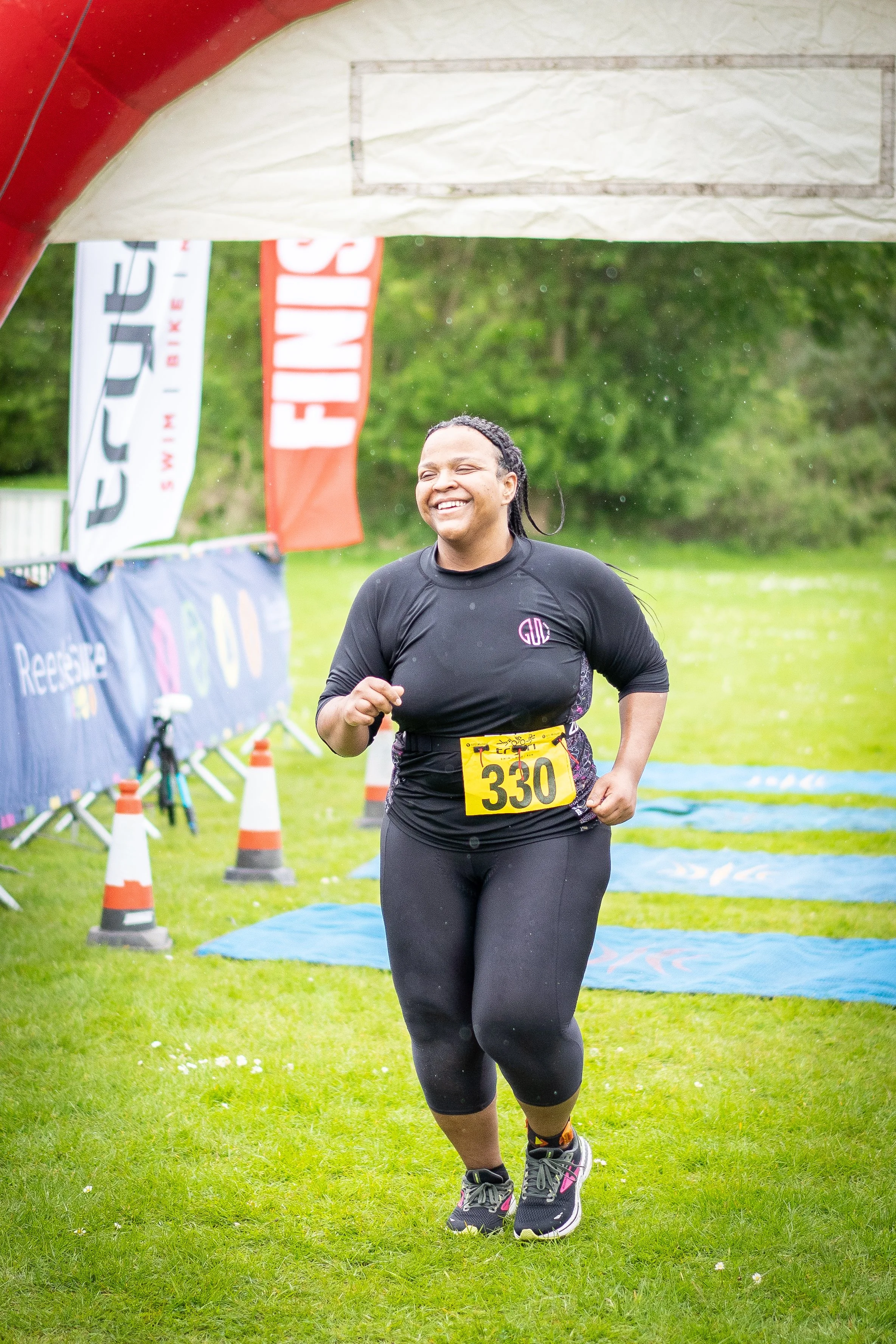 A woman smiling as she crosses the finish line of a race, wearing a black athletic outfit with a yellow bib number 330 in an outdoor park setting.