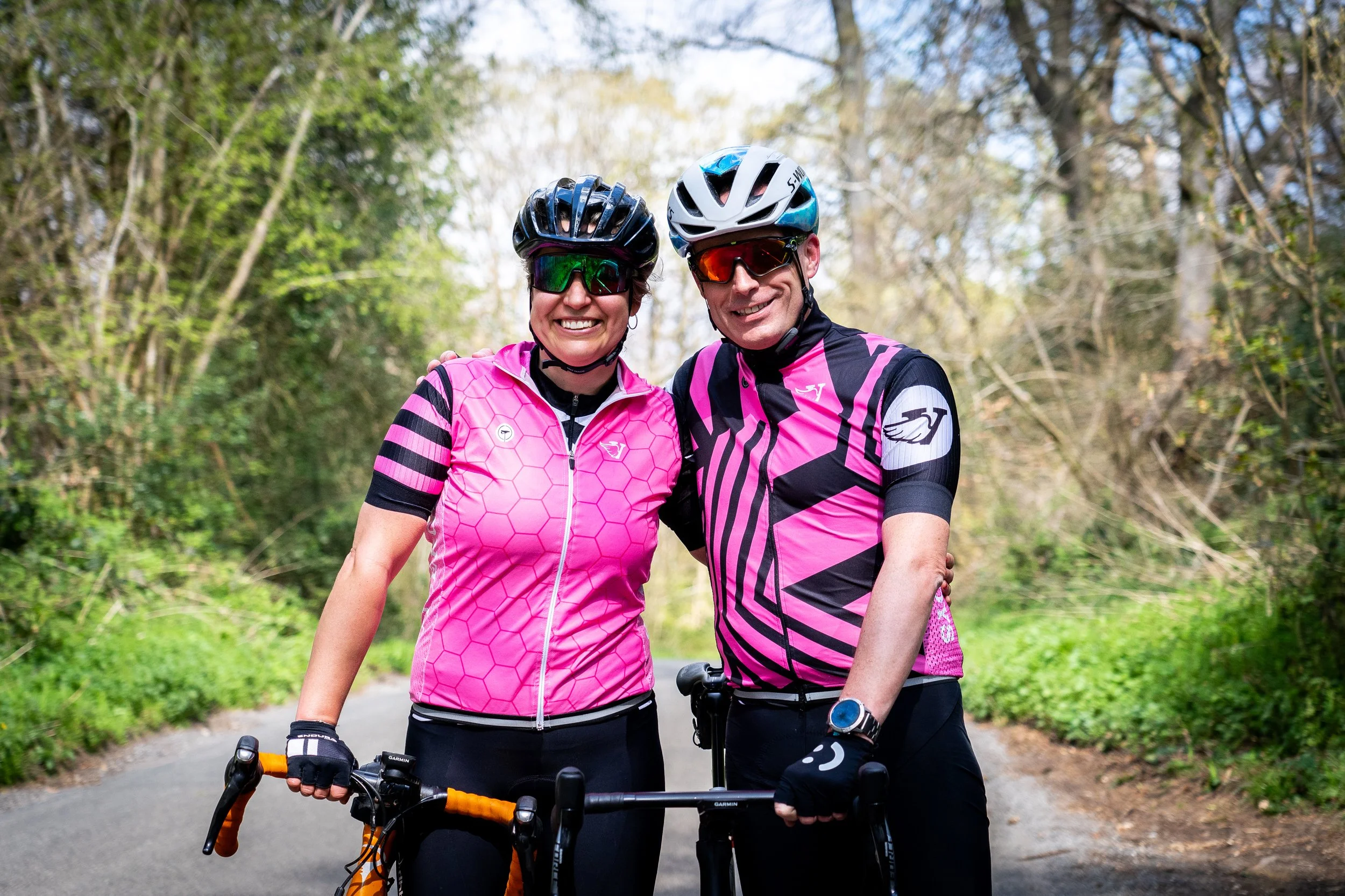 A smiling man and woman in cycling gear and helmets standing outdoors on a wooded path, holding their bikes.