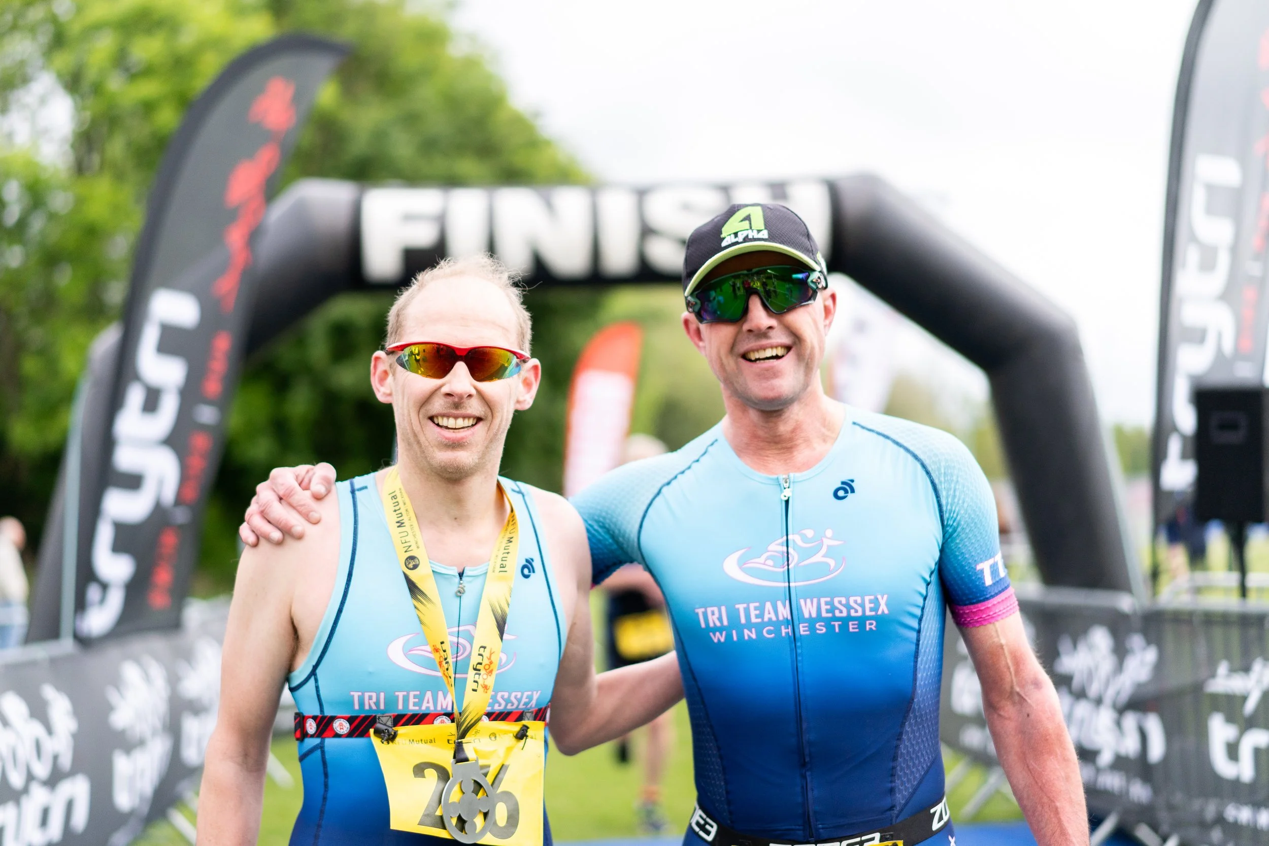 Two smiling male triathletes in blue uniforms at the finish line, wearing sunglasses and medals, with a black and white finish line arch behind them.