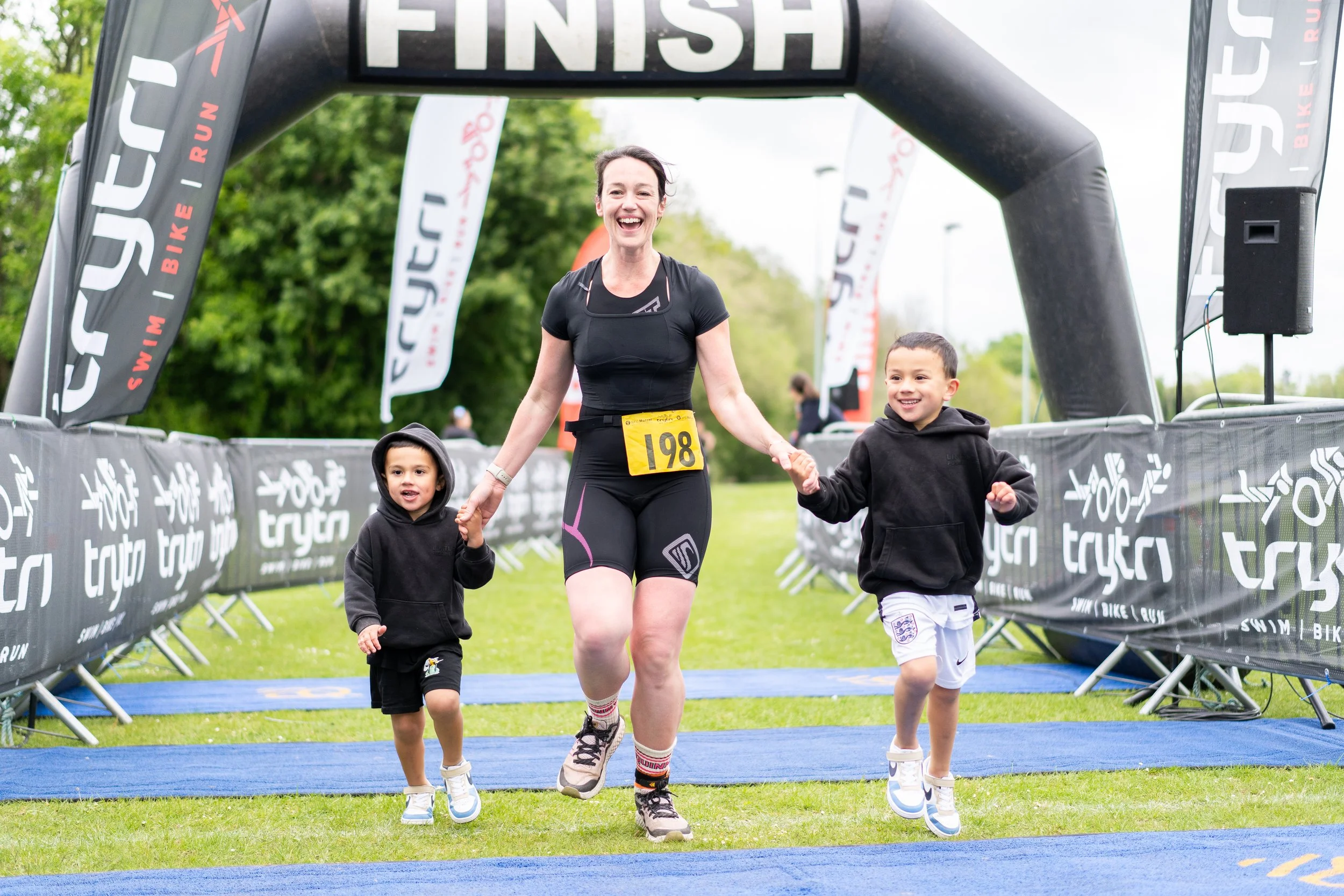 A woman and two young children crossing the finish line of a race, holding hands and smiling. The woman is wearing a black athletic outfit with a yellow race bib numbered 198. The children are wearing black and white hoodies and shorts. The finish li