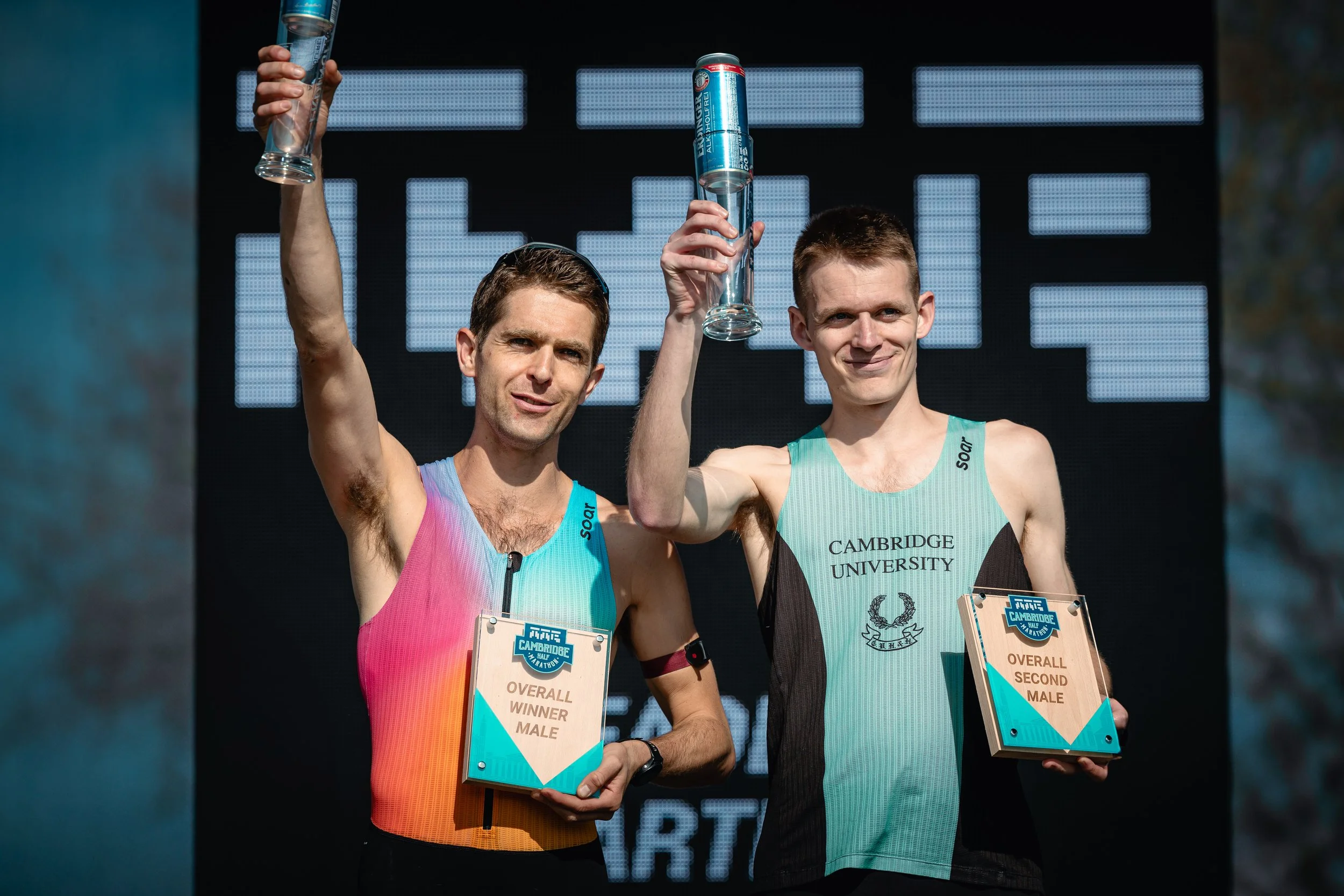Two male athletes in running uniforms holding trophies and glasses, celebrating after a race with a digital scoreboard in the background.