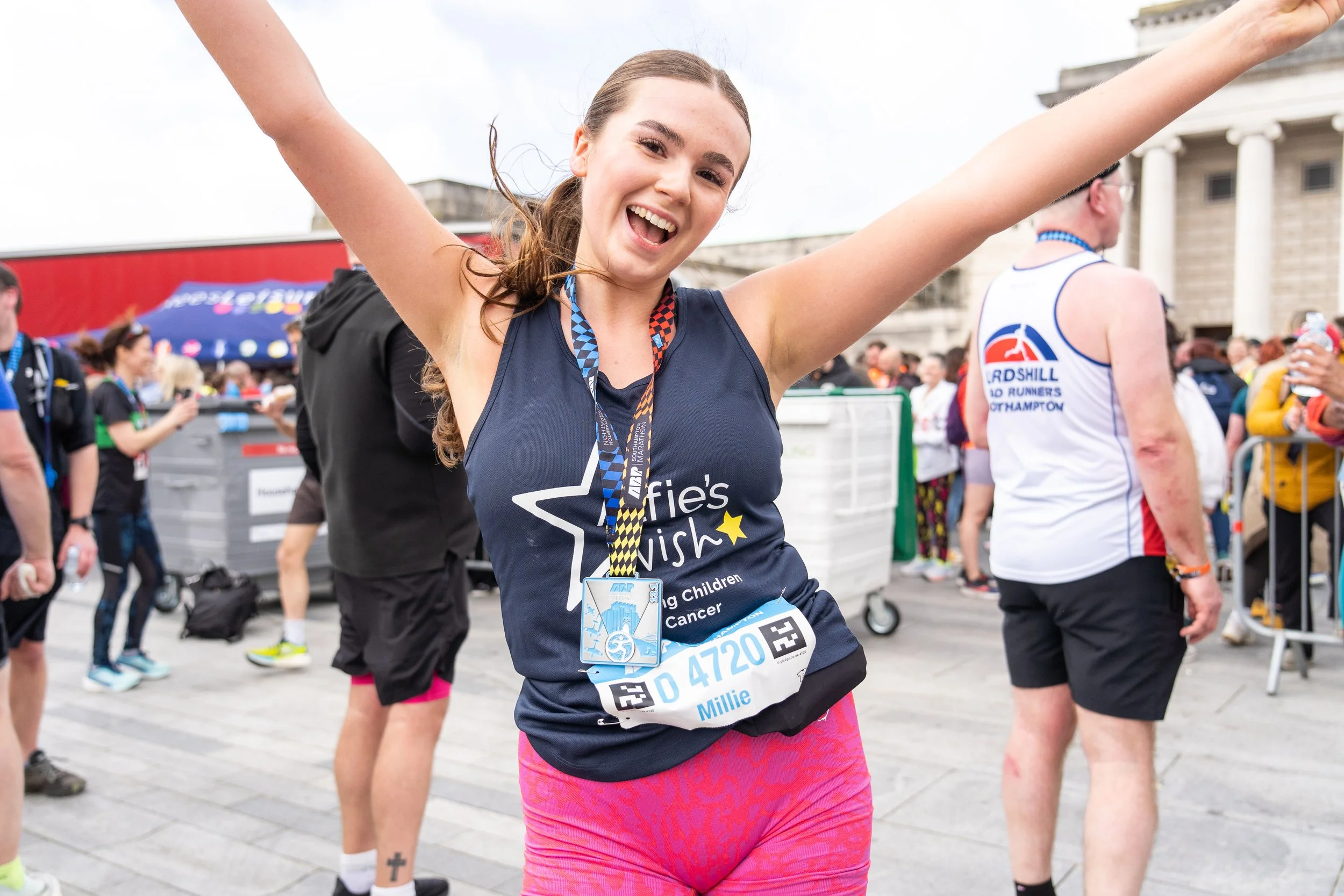 A young woman celebrating after completing a race, wearing a medal and race bib, with her arms raised, in a crowd of runners and spectators outside a large building.