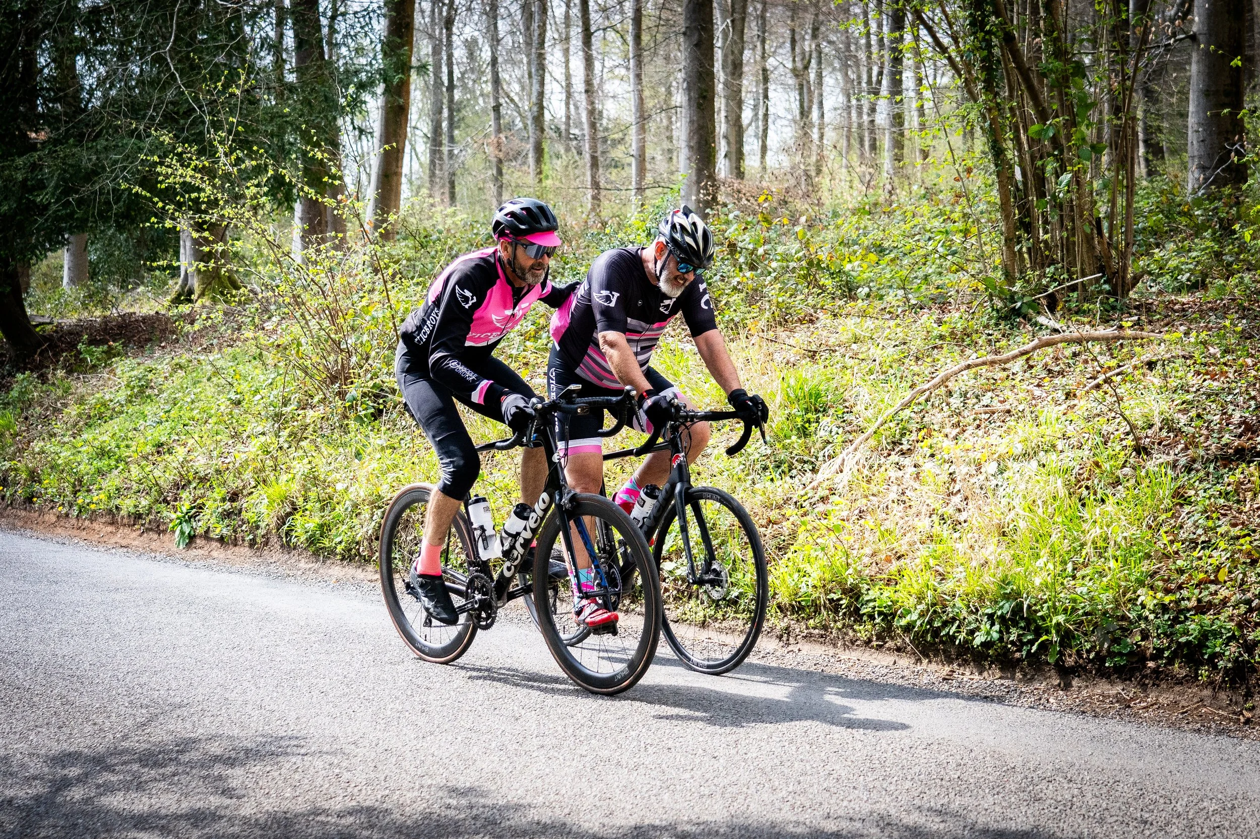 Two men riding bicycles on a forested trail, wearing black and pink cycling outfits and helmets.