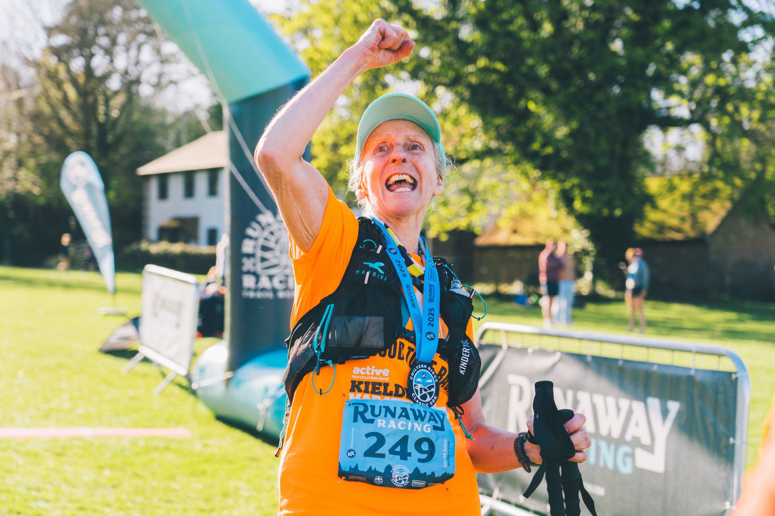 An elderly woman celebrating at the finish line of a race, wearing an orange shirt, a race bib numbered 249, a medal, and a cap, with a triumphant expression and her fist raised.