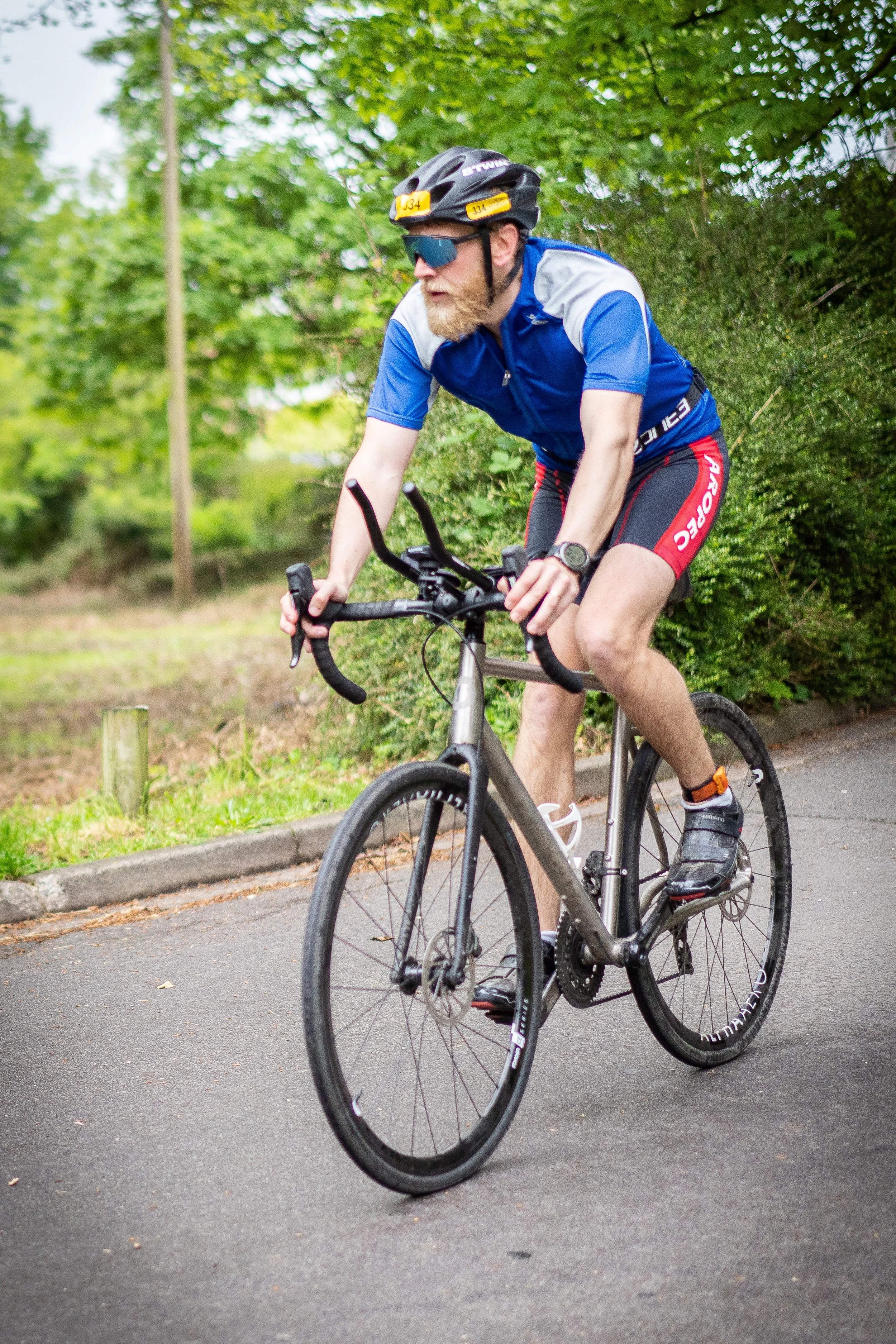 A man with a beard biking on a paved path surrounded by green trees, wearing a helmet, sunglasses, a blue athletic shirt, red and black shorts, and cycling shoes.