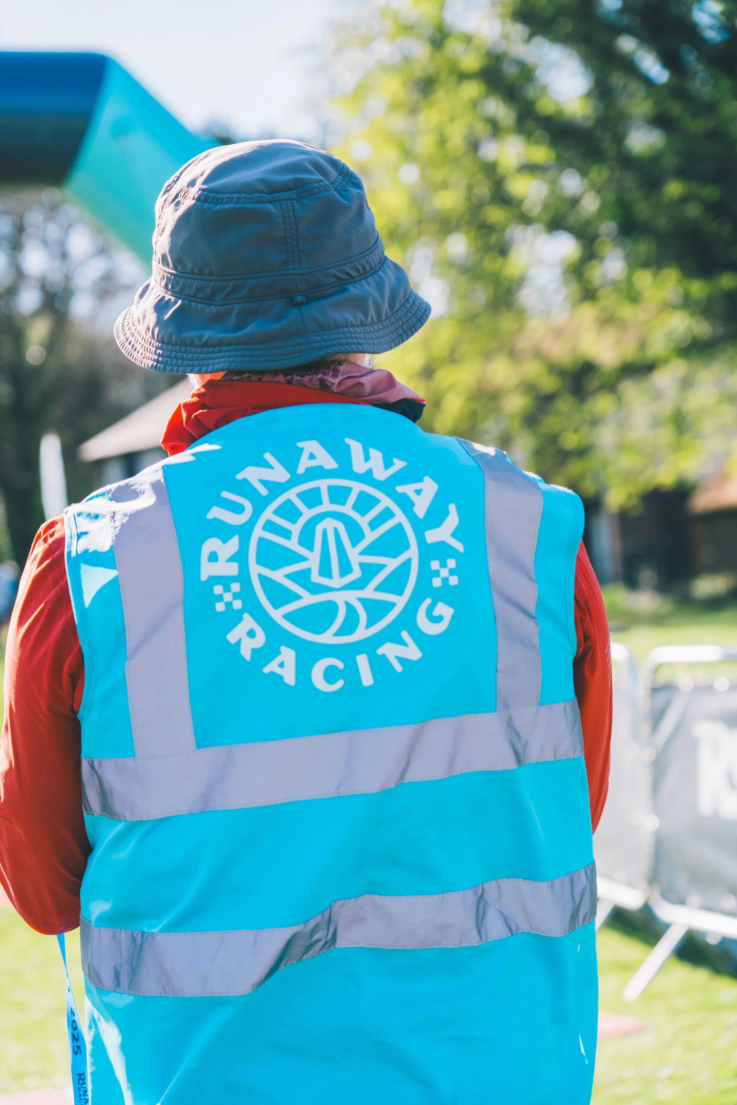 Back of a person wearing a blue vest with 'Trail Run Day Racing' logo, red long-sleeve shirt, gray bucket hat, and a red and purple neck gaiter, standing outdoors with trees in background.