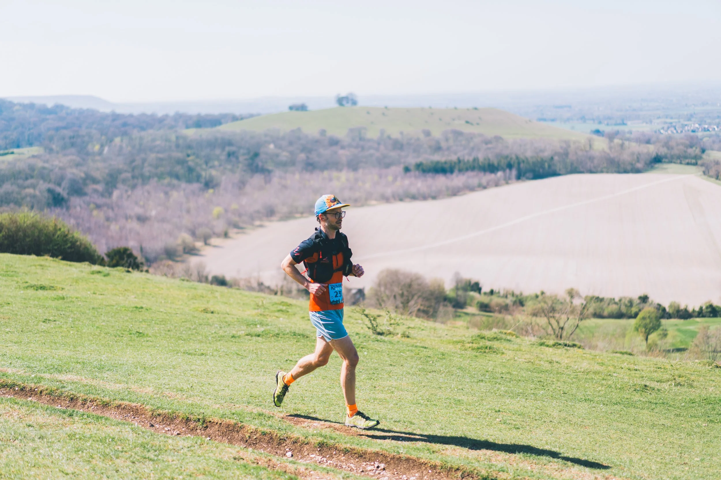 A man running on a grassy trail in a hilly landscape with fields and trees in the background.