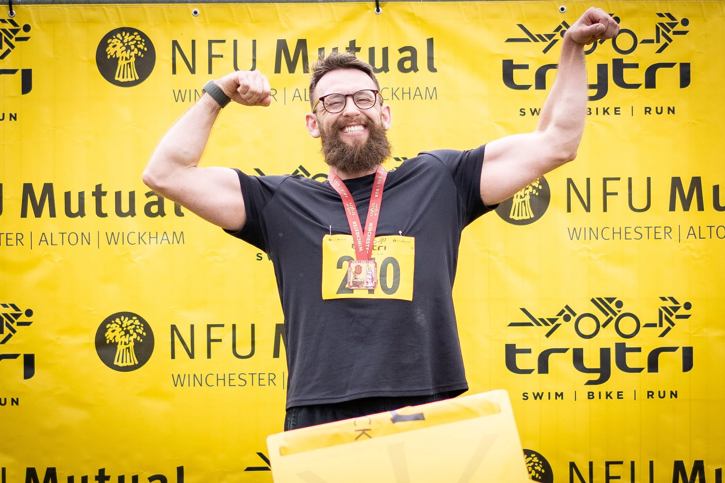 A man with a beard and glasses, wearing a black shirt, flexing his muscles and smiling at a sports event, with a yellow backdrop behind him featuring the NFU Mutual and TryTri logos.
