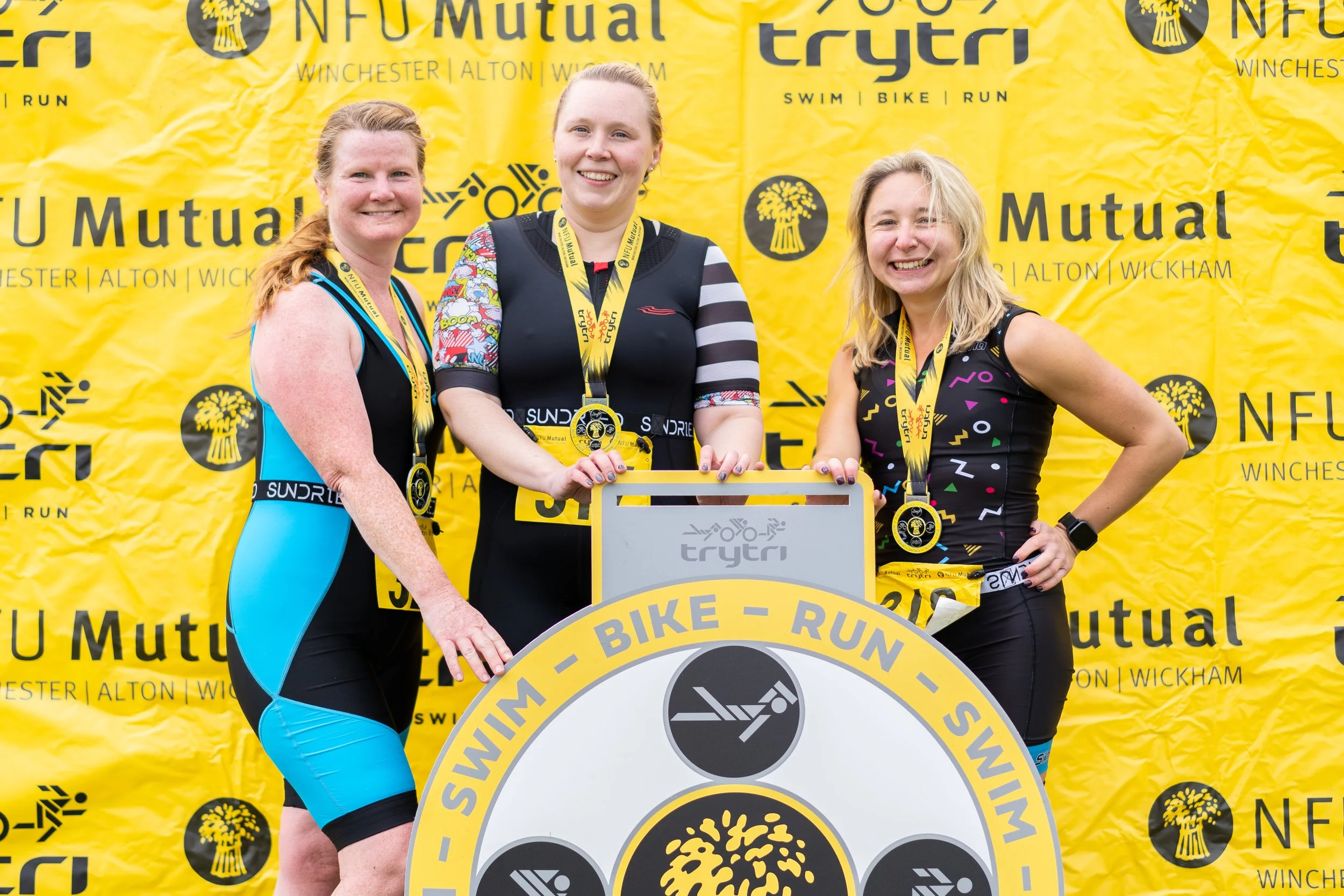 Three women in athletic gear wearing medals contesting in a triathlon finish line, holding a large circular sign with Triathlon icons, in front of a yellow backdrop featuring "NFU Mutual" logos and mountain drawings.