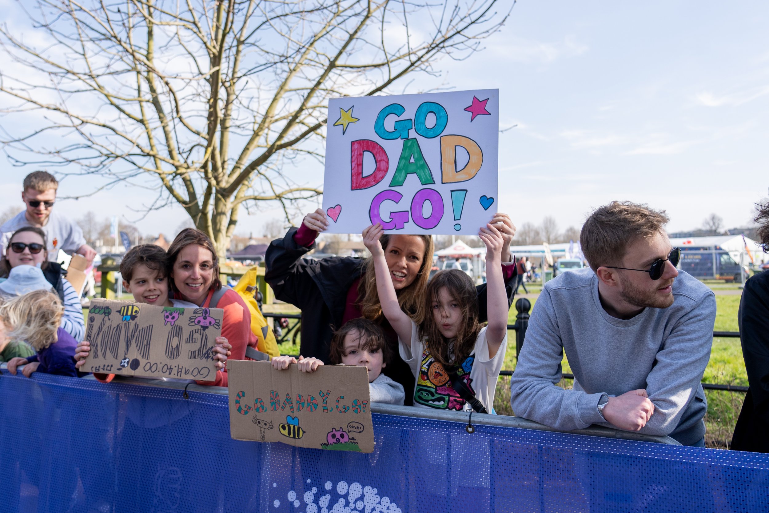 People gathered outdoors at a festival, holding colorful hand-drawn signs including one that says "GO DAD GO!" in large letters. The group includes children and adults standing behind a blue barrier, some smiling and some posing with the signs.