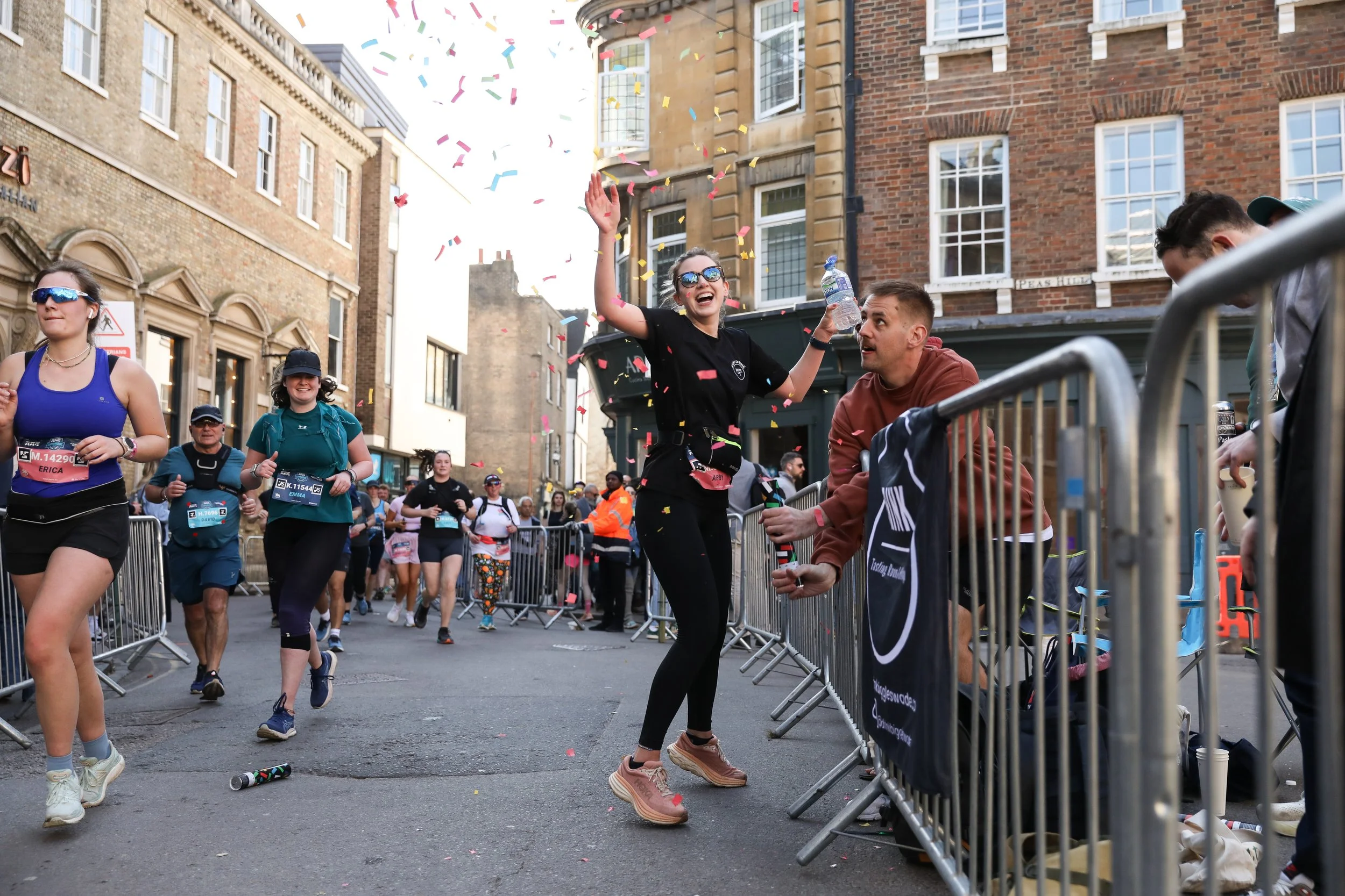 People participating in a marathon or race, with some celebrating near a barrier and others running behind, in an urban street setting with old brick and stone buildings. Confetti is falling, and a woman in black with sunglasses is smiling and raisin