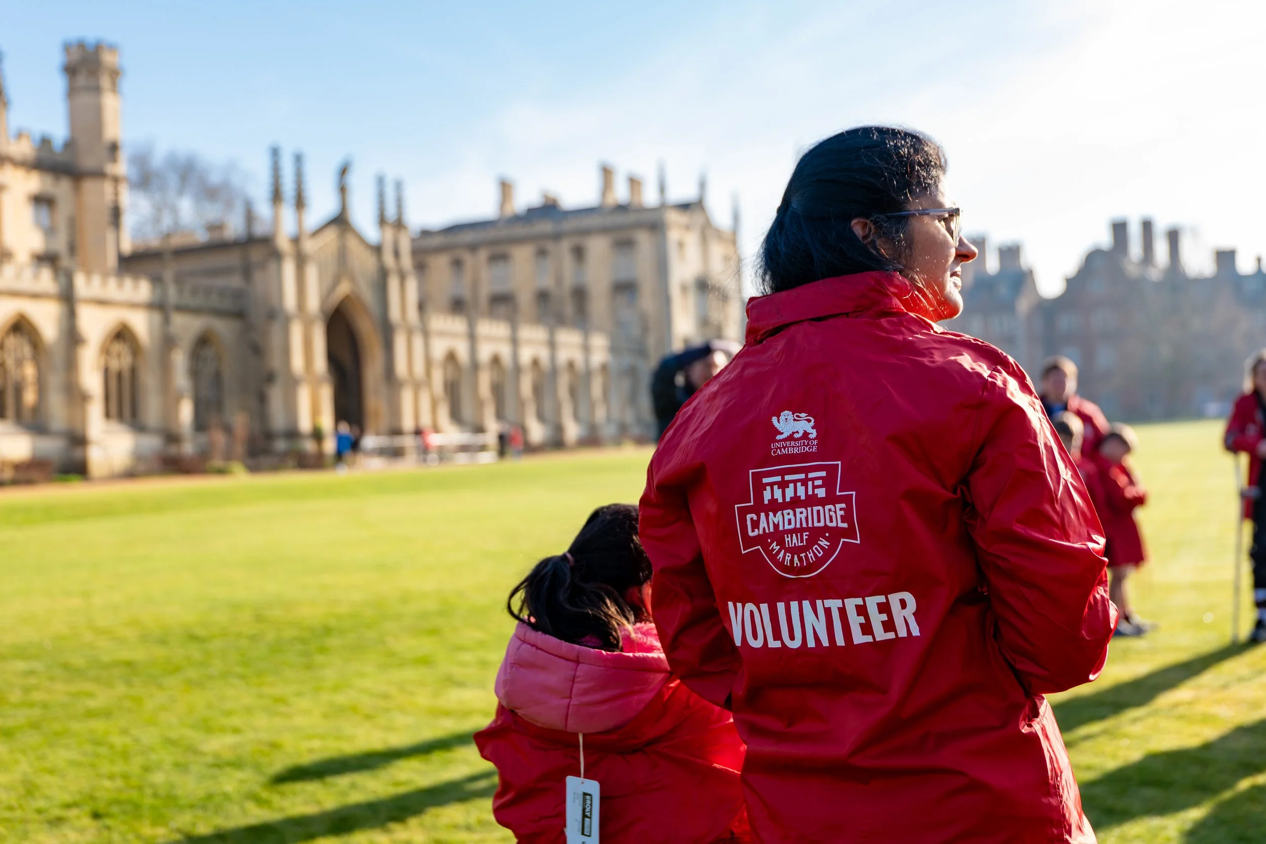 A woman wearing a red volunteer jacket with 'Cambridge Half Marathon' logo stands on a grassy field during a daytime outdoor event, with a historic building in the background.