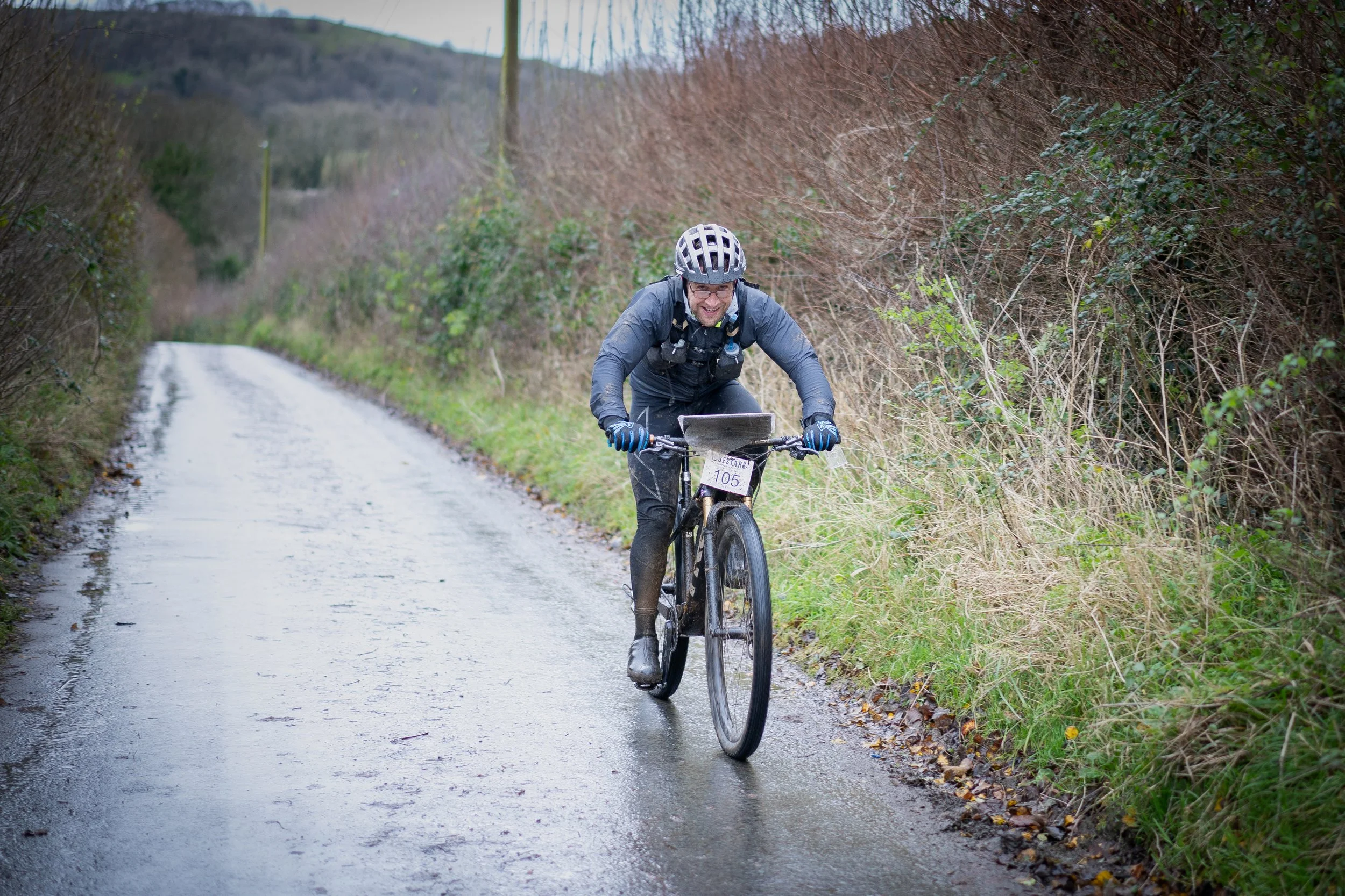Man riding a mountain bike on a wet, rural trail during overcast weather, wearing a helmet and outdoor gear.