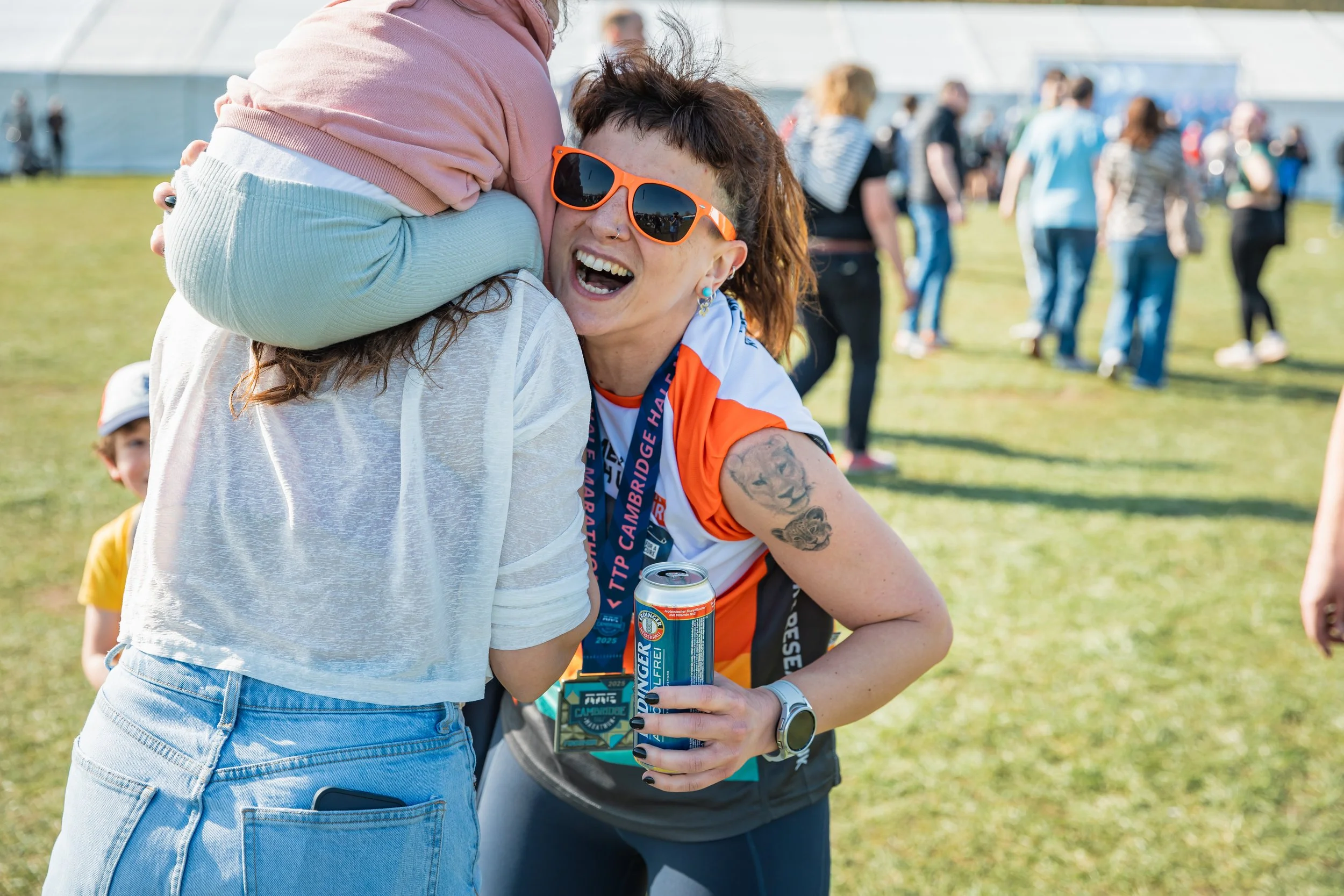 Two women hugging and smiling at an outdoor event on a sunny day, with a crowd of people in the background.