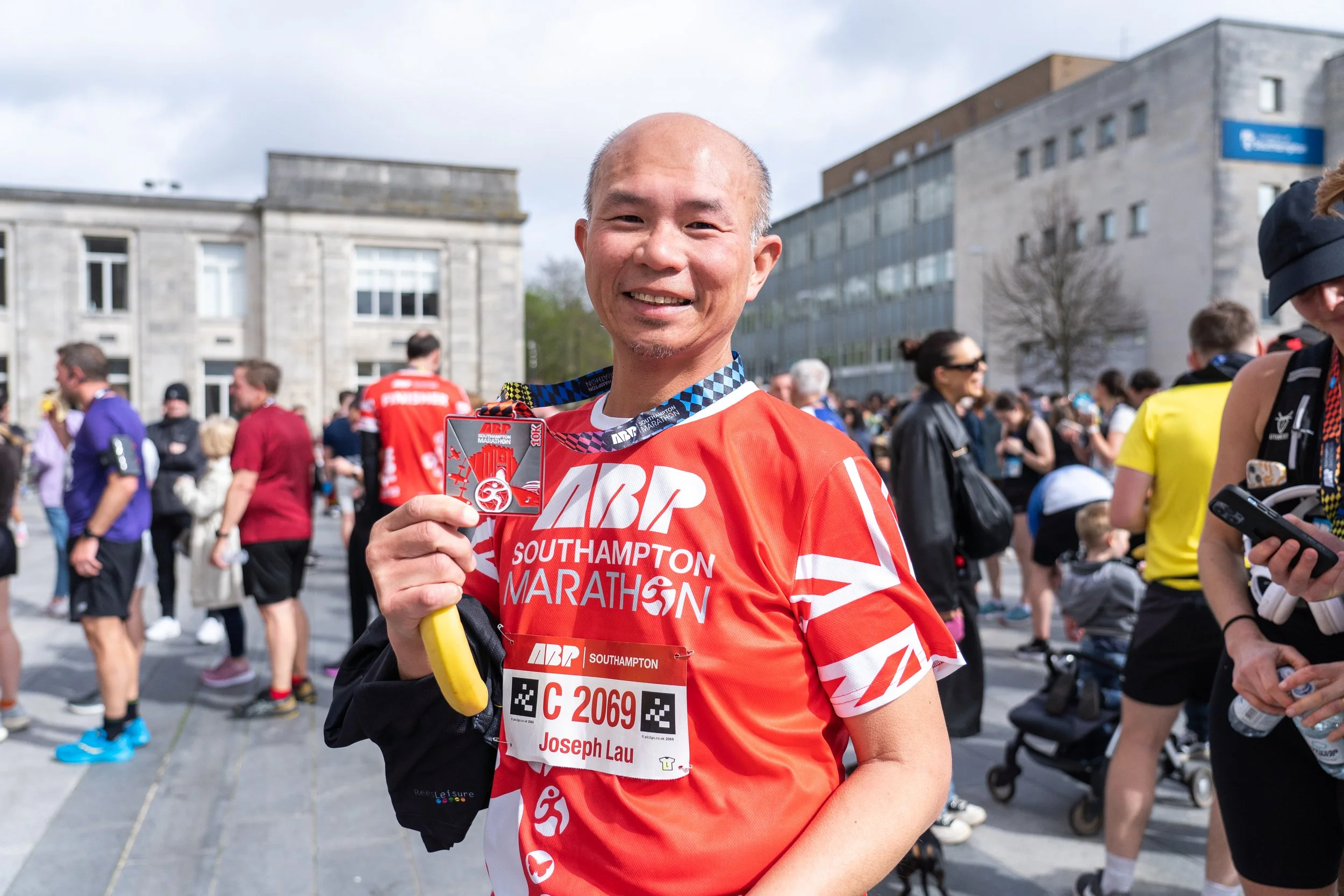 Smiling male marathon runner in red shirt holding a medal at a marathon event, with a crowd of people in the background