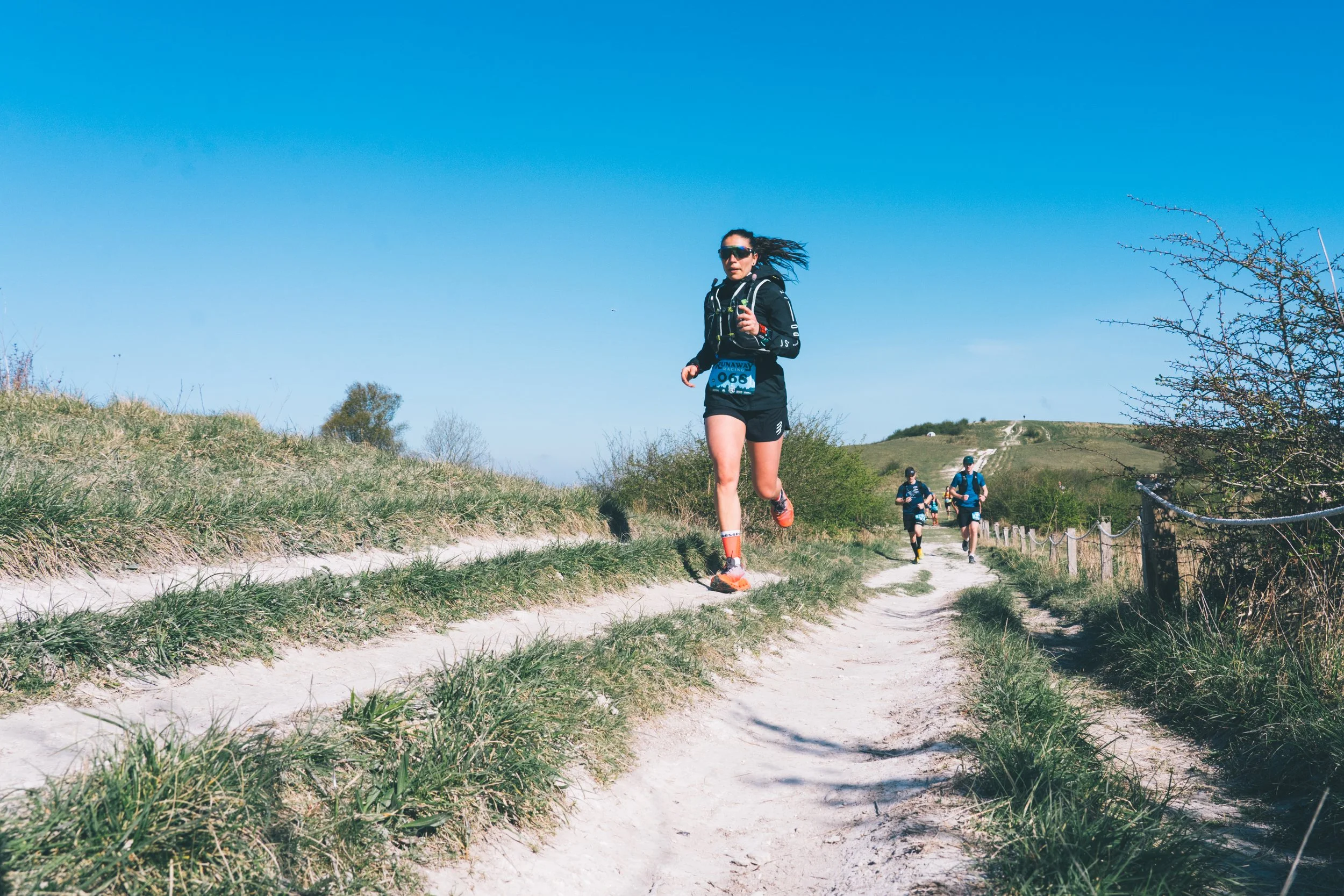 A female runner in black sportswear and sunglasses running on a dirt trail in nature with three other runners in the background on a sunny day