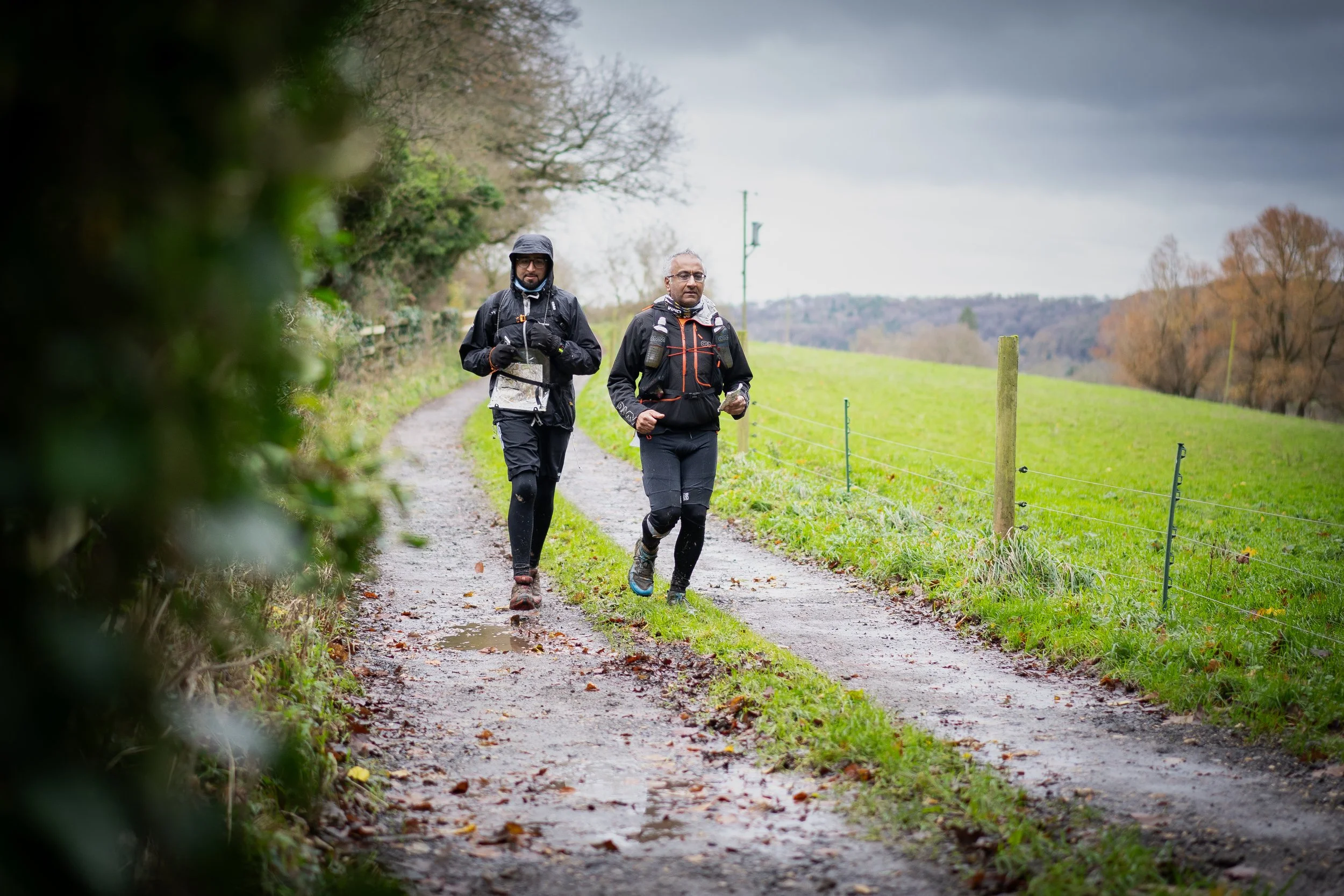 Two men hiking on a muddy trail through a countryside landscape with green grass and leafless trees, overcast sky in the background.