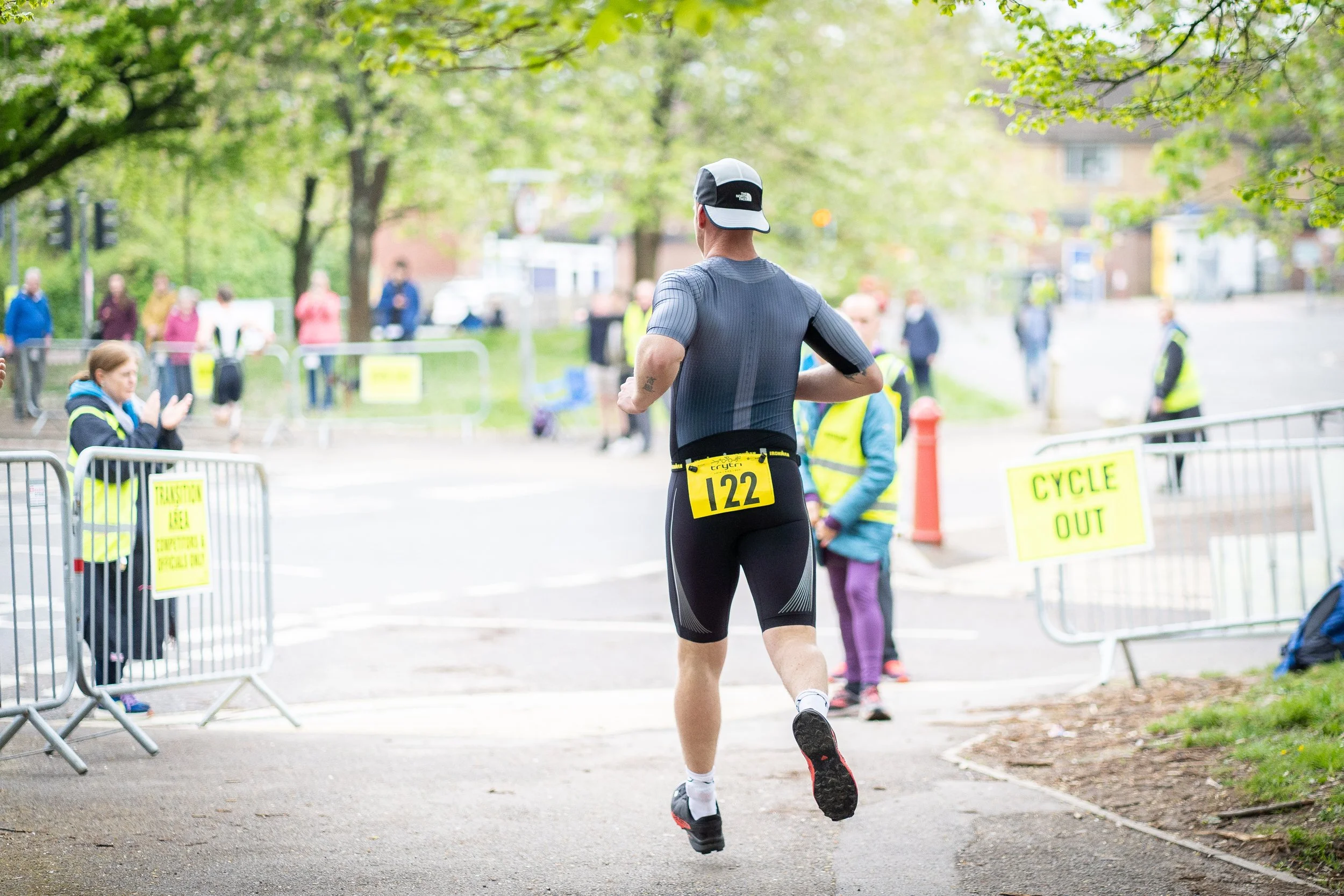 Male athlete running in a race, wearing a gray athletic shirt, black shorts, and a white cap, with race number 122 attached to his waist, in a park with volunteers and signs indicating race directions.