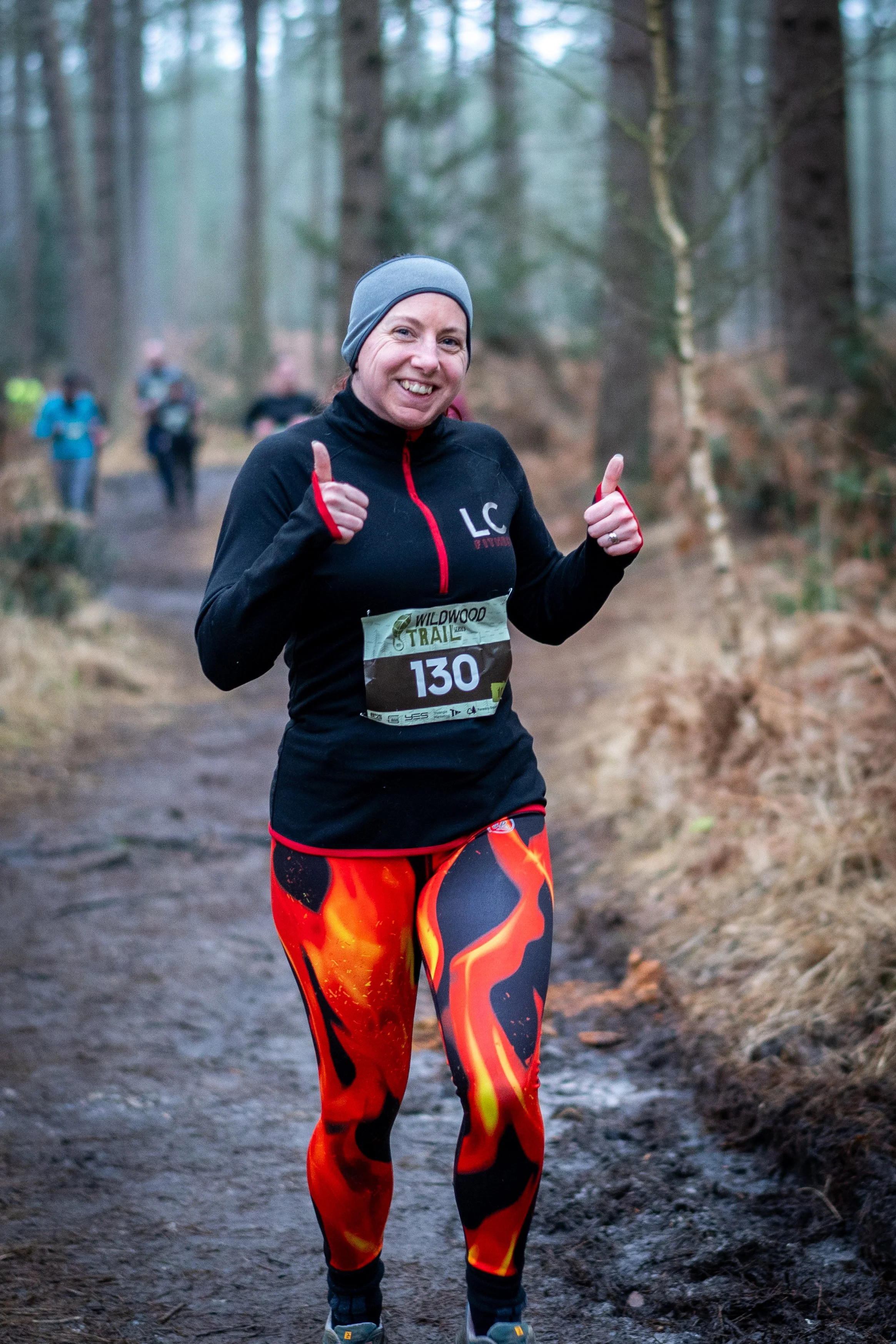 A woman in athletic gear giving a thumbs-up during a trail run in a forest.