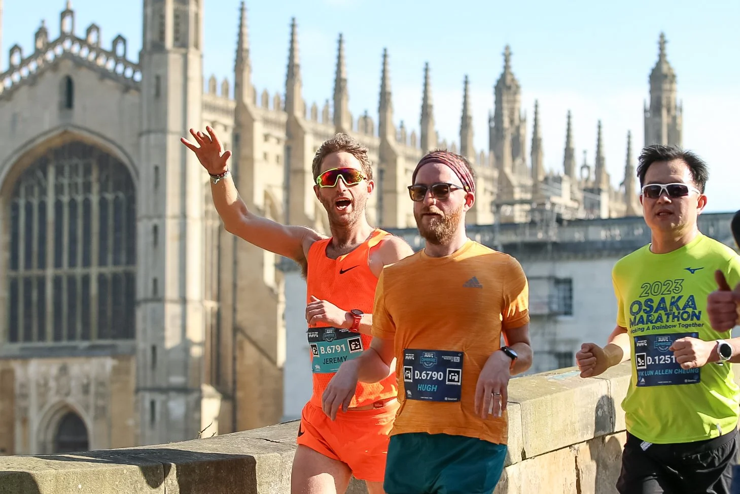 Three male marathon runners in athletic gear running past a historic stone building with tall spires, during daytime.