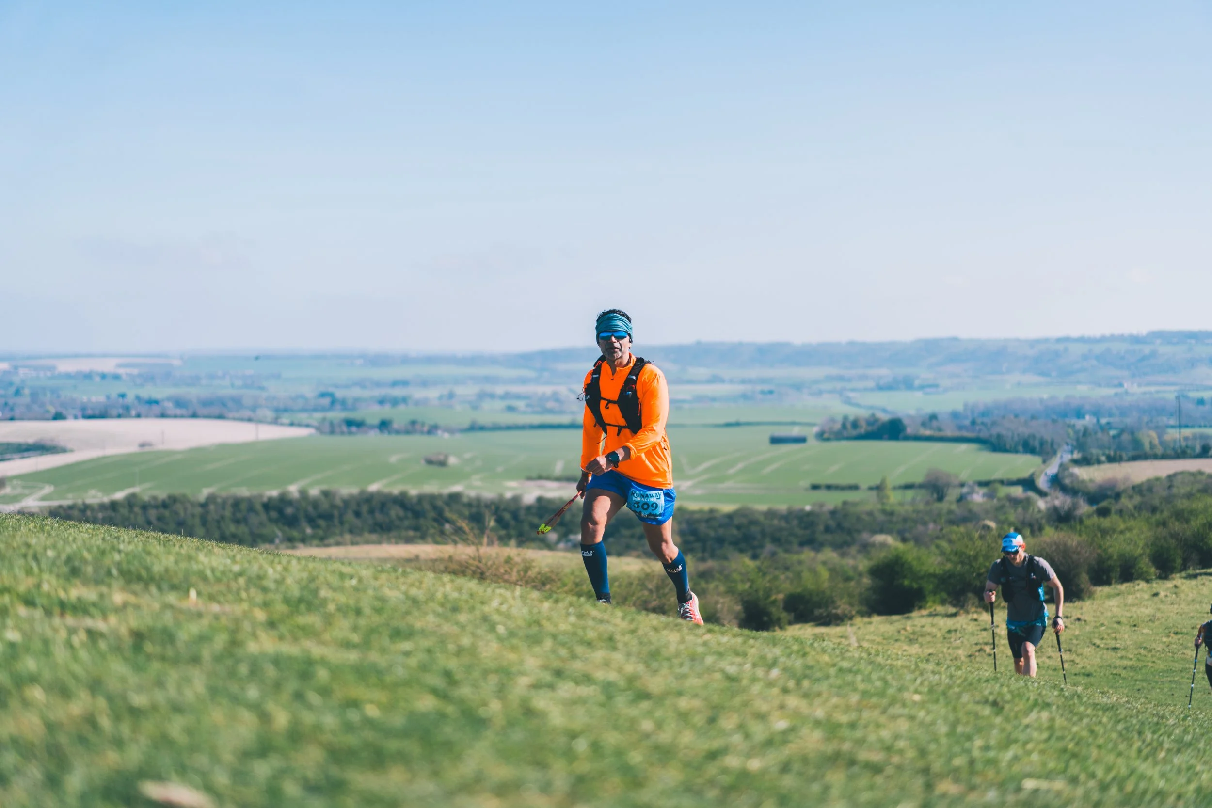 Two runners participating in a trail race on a grassy hillside with rolling green fields and trees in the background under a clear blue sky.
