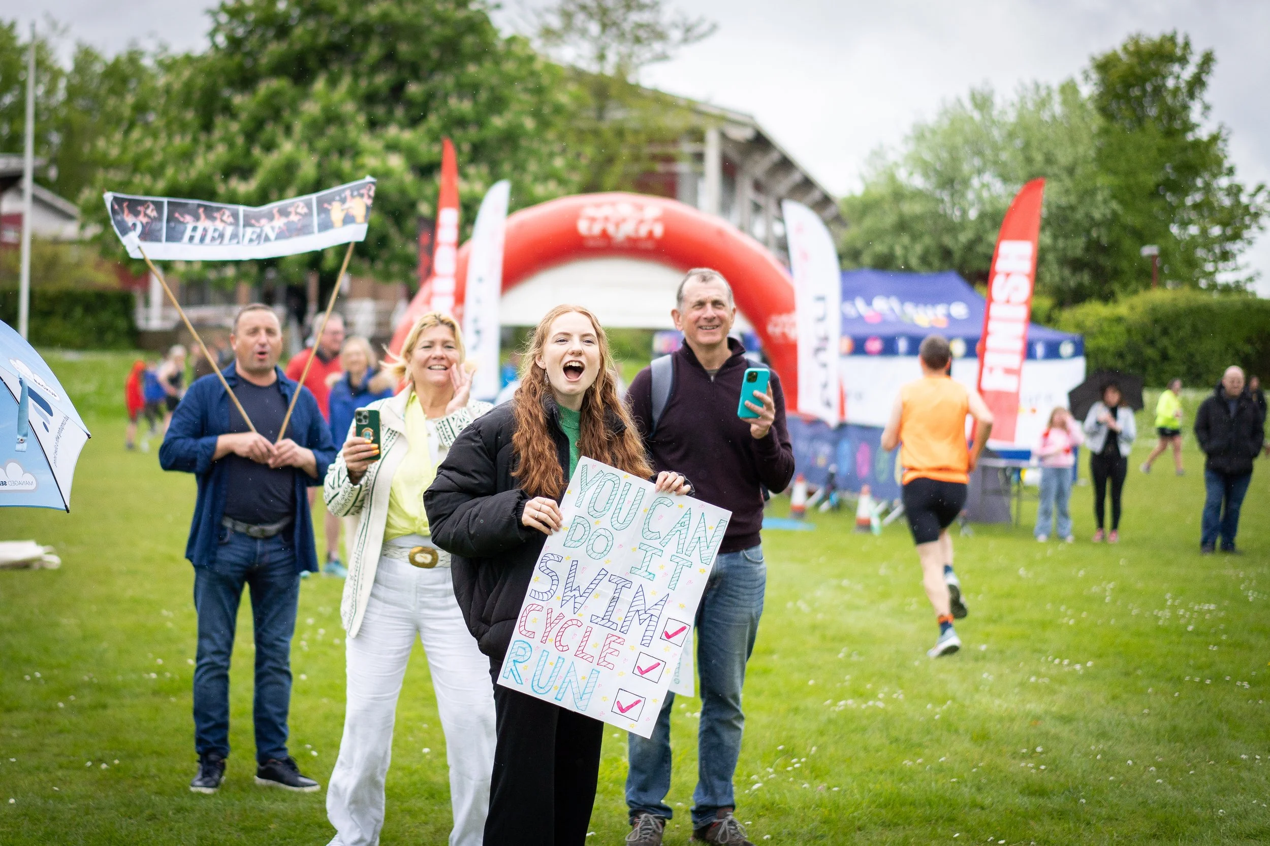 Group of people cheering at an outdoor event, with a woman holding a colorful sign that says "You Can Do It! Swim, Cycle, Run" and others taking photos or holding umbrellas.