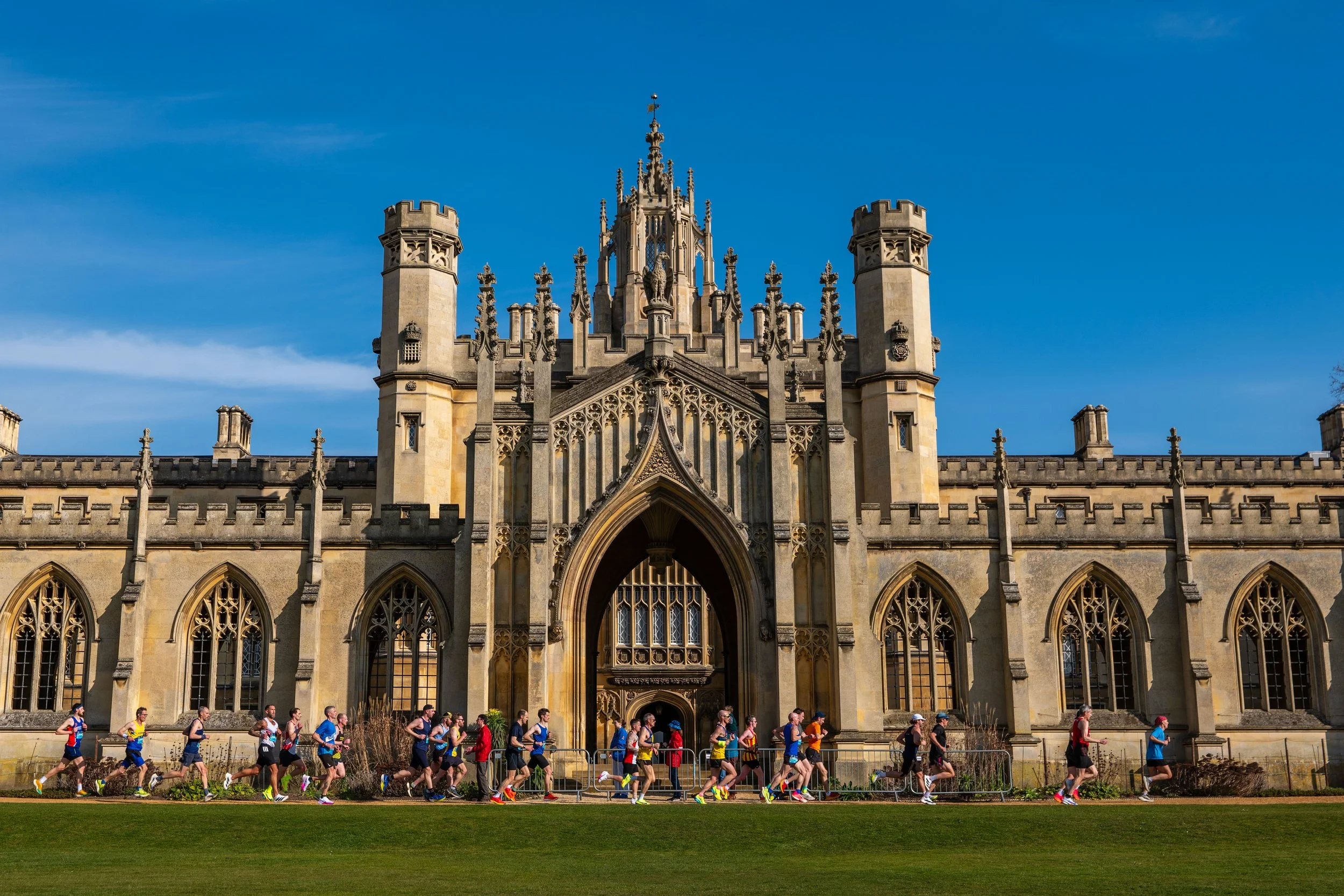Marathon runners passing in front of a historic Gothic-style building with towers, arched windows, and ornate stonework, under a clear blue sky.