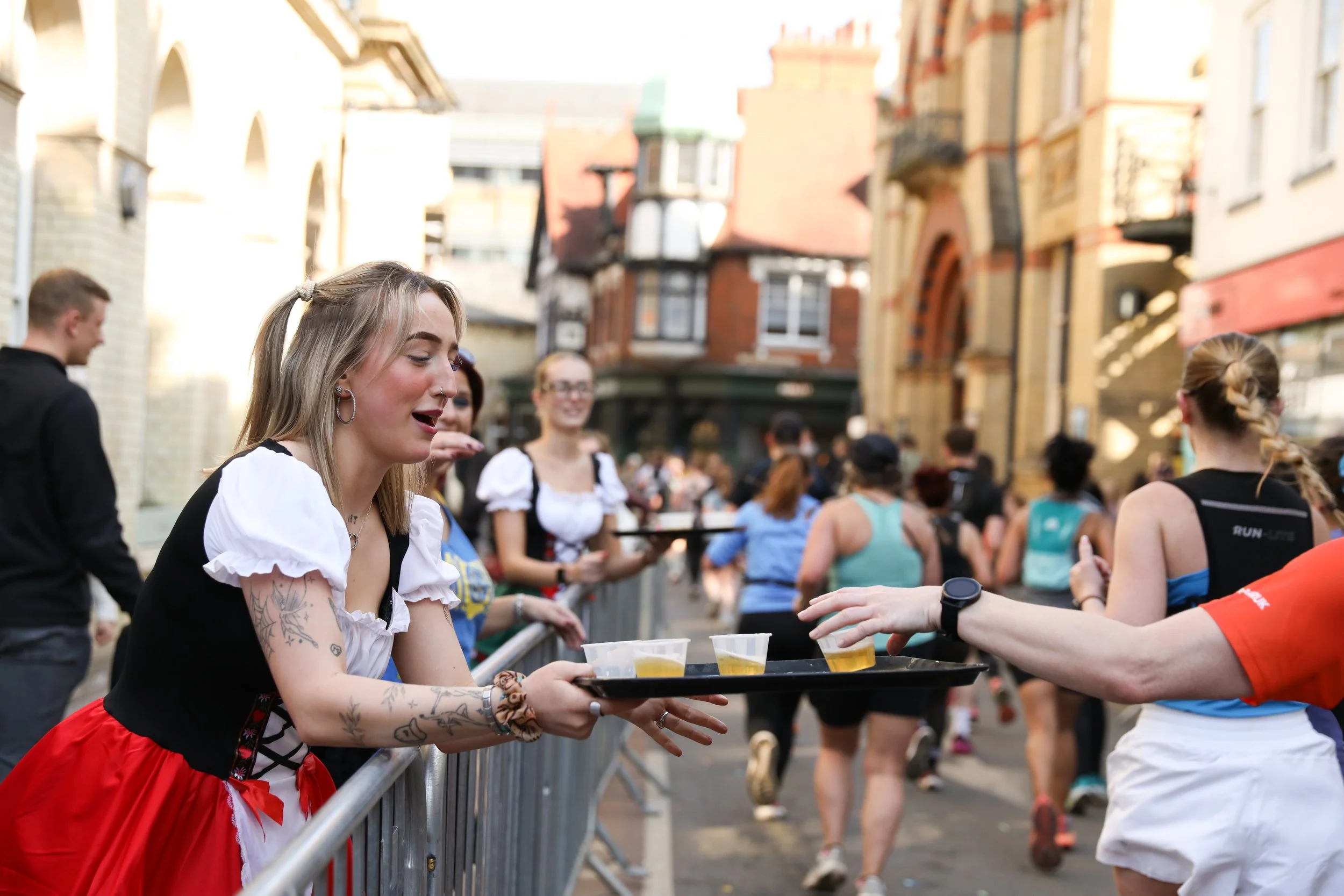 A woman in a traditional dress handing out cups of beer to runners at a race event on a city street.