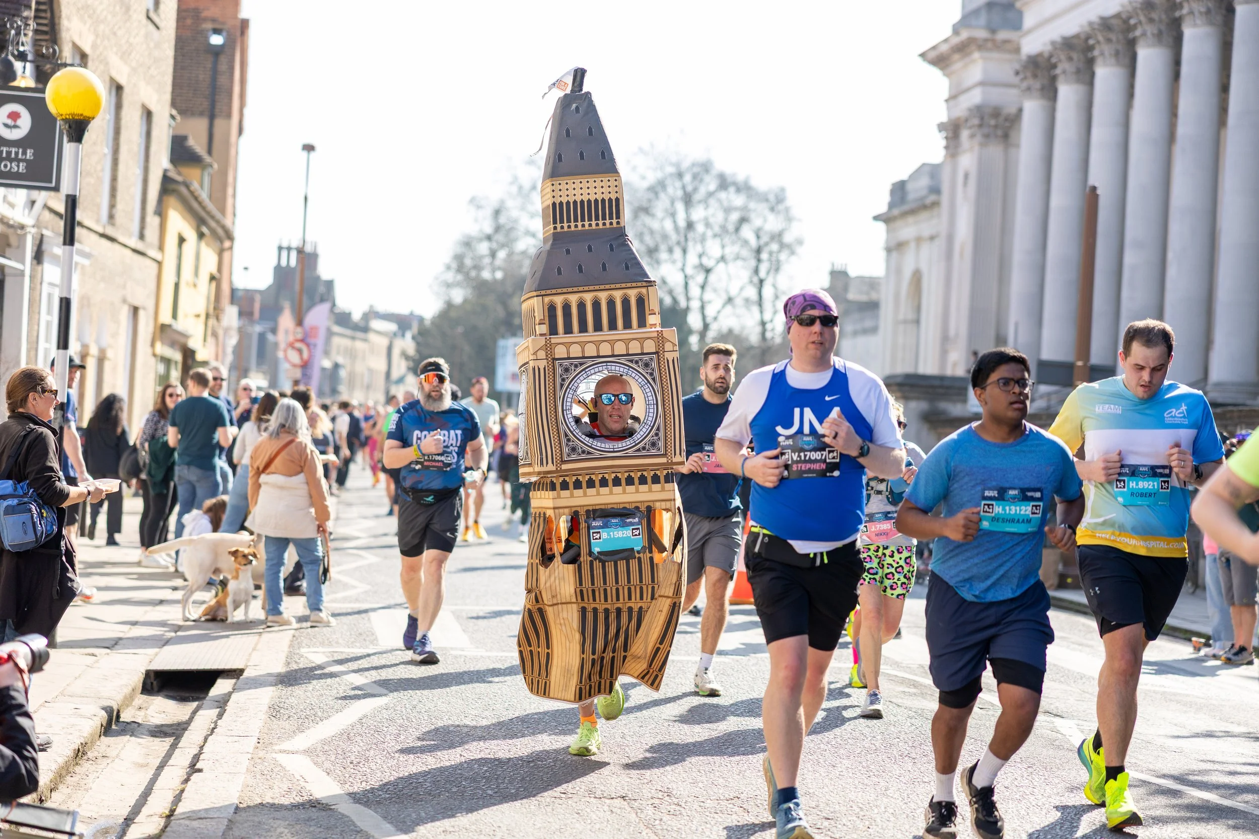 Group of marathon runners participating in a race, with one runner dressed in a Big Ben clock tower costume, running along a street lined with spectators and historic buildings on a sunny day.