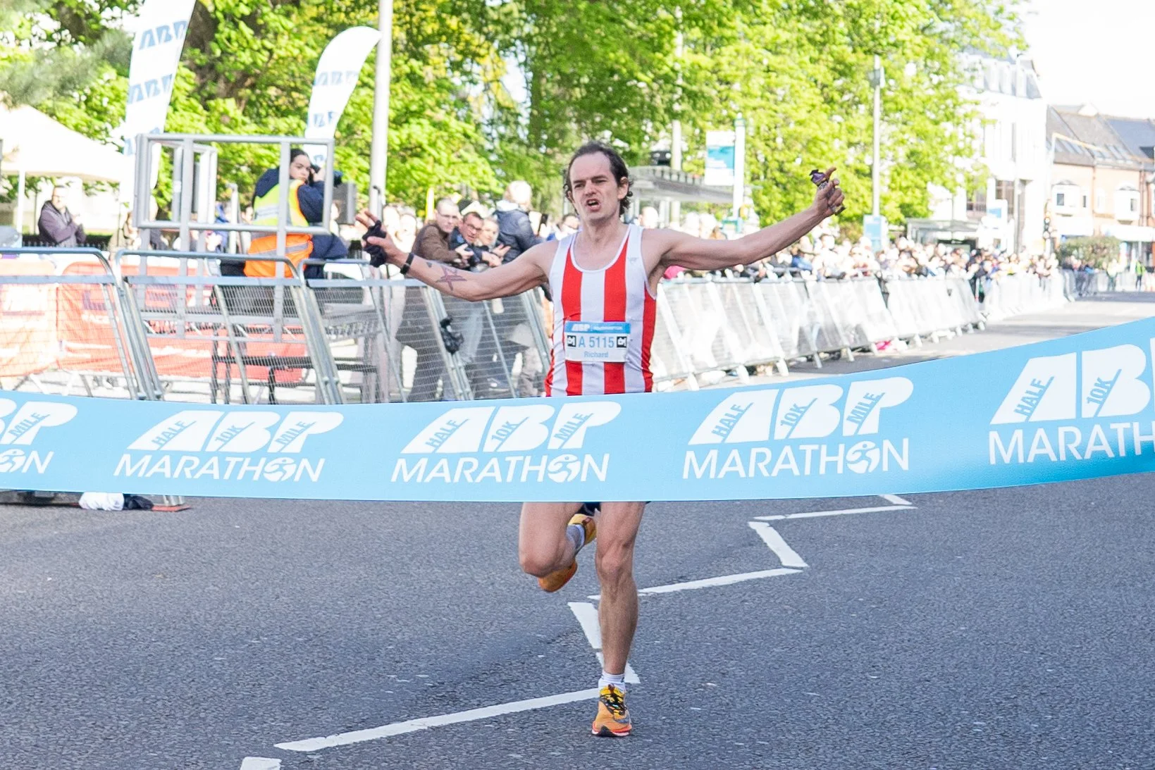 Runner crossing the finish line at the Half Marathon, wearing a red and white striped tank top, with arms raised in victory, and crowd cheering in the background.