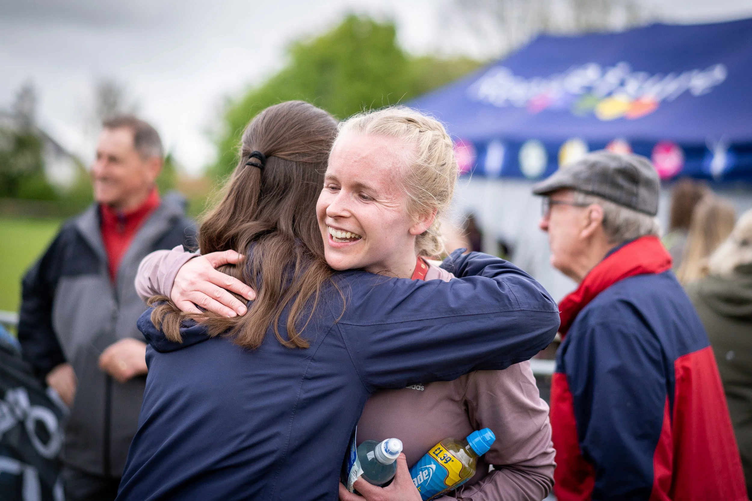 Two women hugging each other, smiling, at an outdoor event with a blue tent in the background. Other people are visible in the background, some wearing jackets.