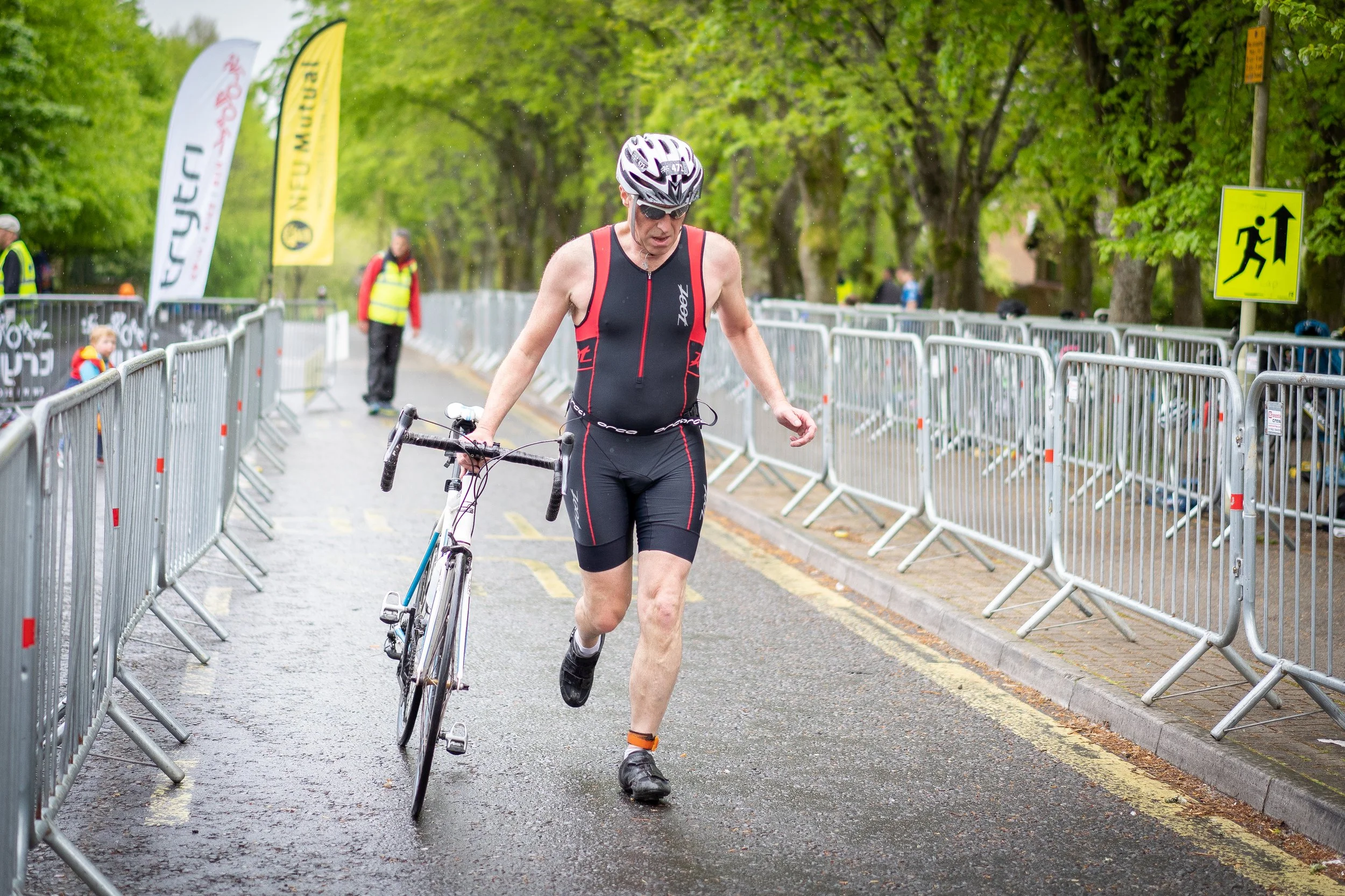 Male triathlete in black and red athletic gear, wearing a helmet and sunglasses, runs on wet pavement while carrying a bicycle during a race event, with metal barriers lining the street and trees in the background.