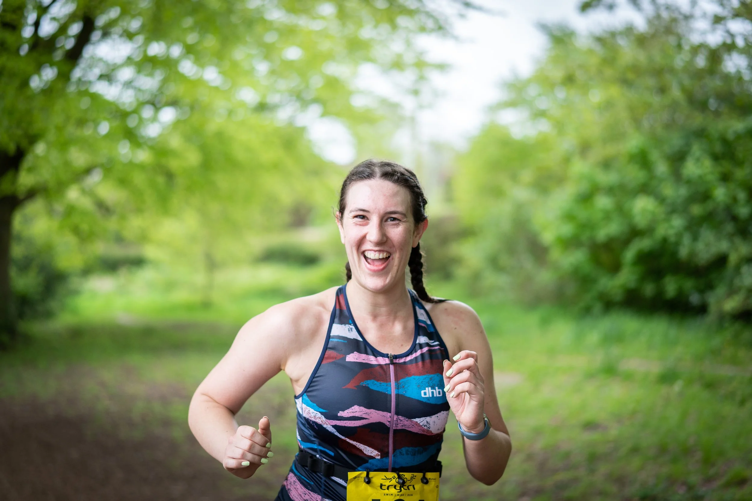 A young woman jogging outdoors on a forest trail, smiling and wearing athletic gear and a yellow race bib.