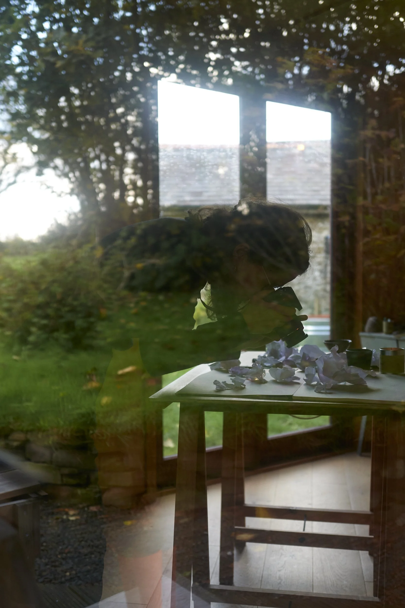 Reflection of a woman with glasses taking a photograph through a glass door or window, with a table and crumpled paper in foreground and greenery and windows in the background.