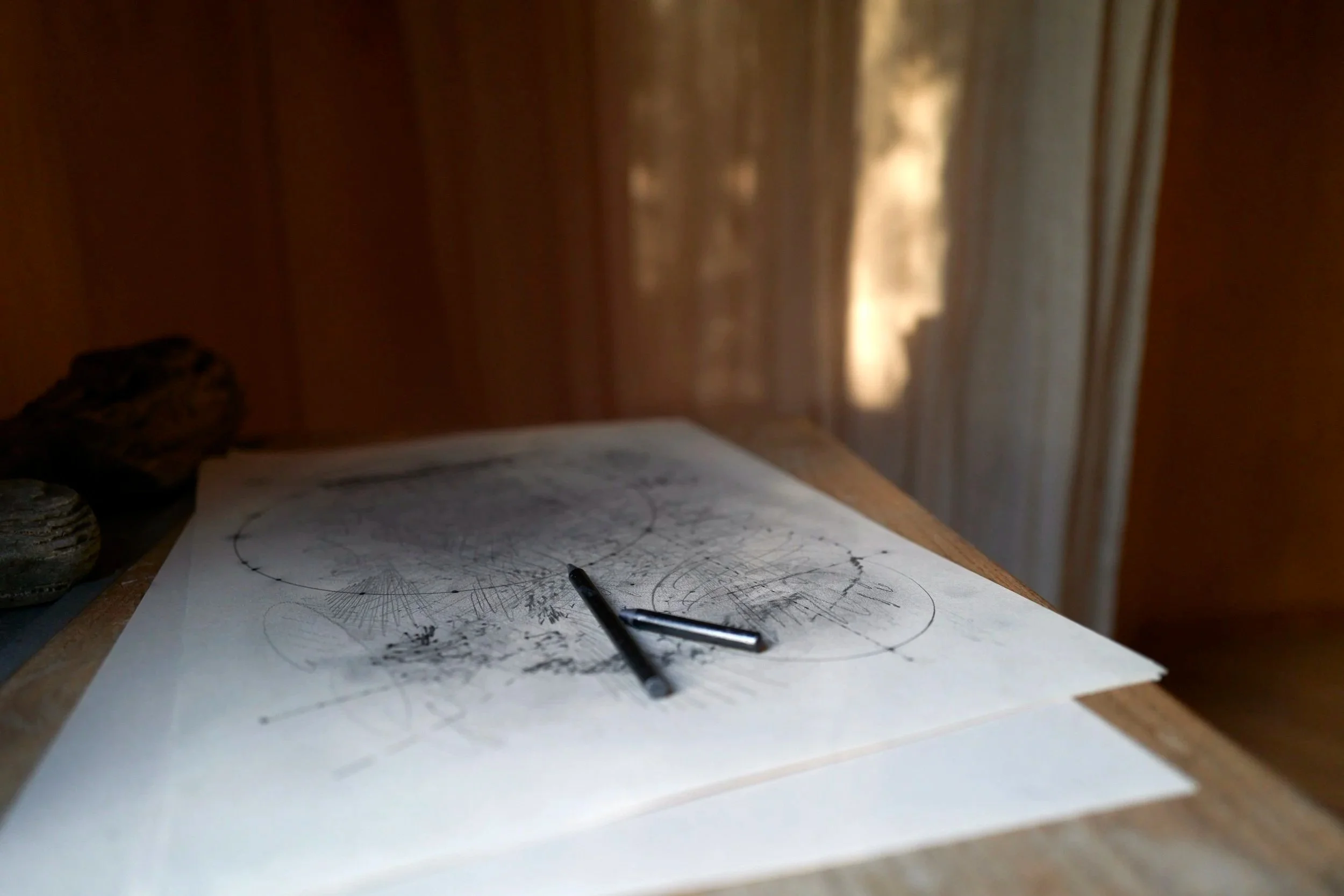 A wooden desk with a sketchpad featuring abstract geometric drawings, two black pens, and some rocks, with sunlight coming through curtains in the background.