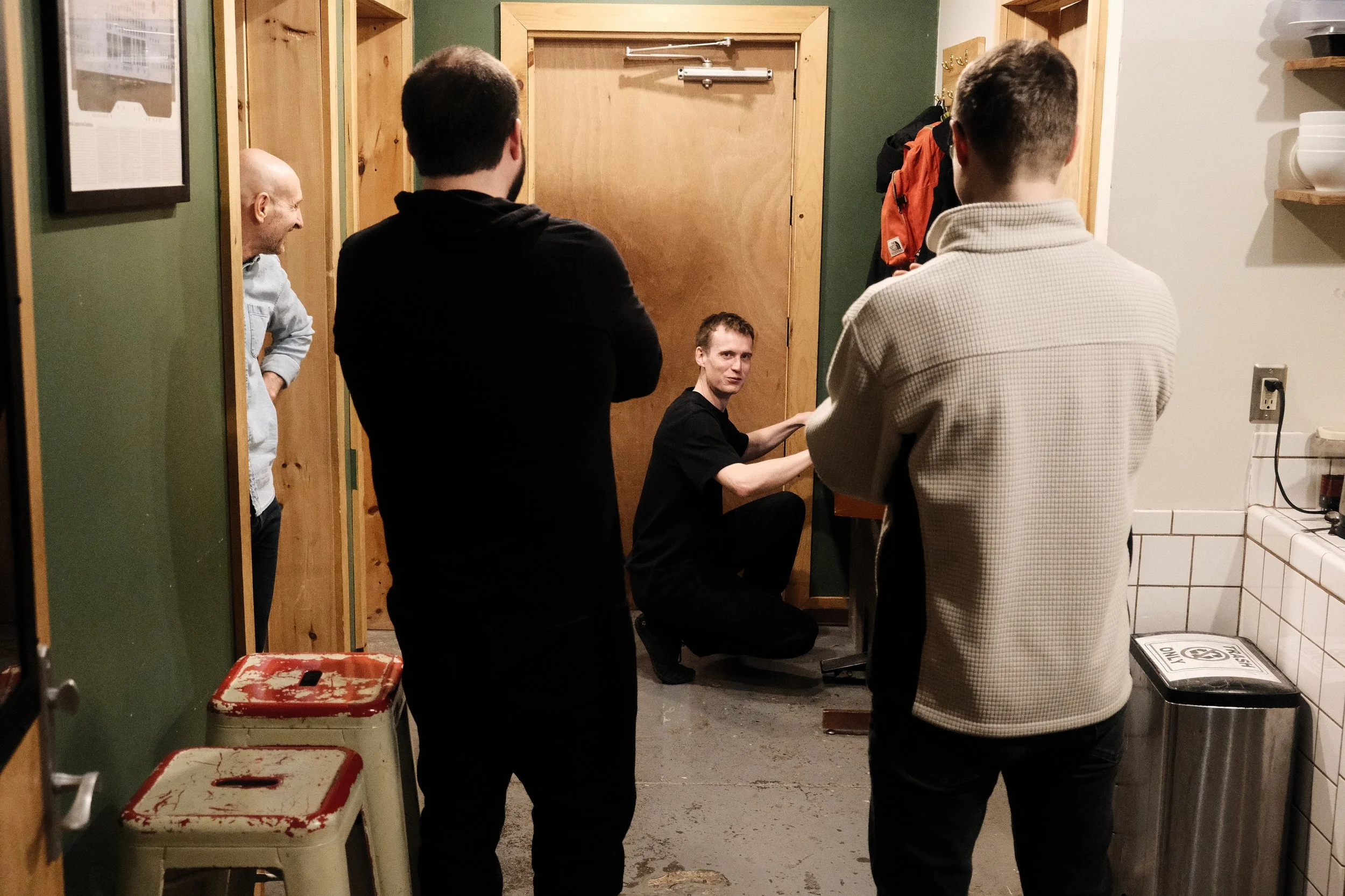 Group of men in hallway, one crouching by wooden door, others standing, some stools, green and white walls.