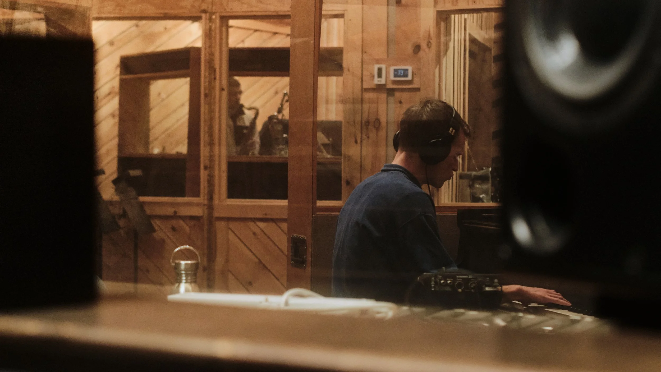 Musician in recording studio playing piano, wearing headphones, with sound equipment and saxophonist visible in background, wooden walls.